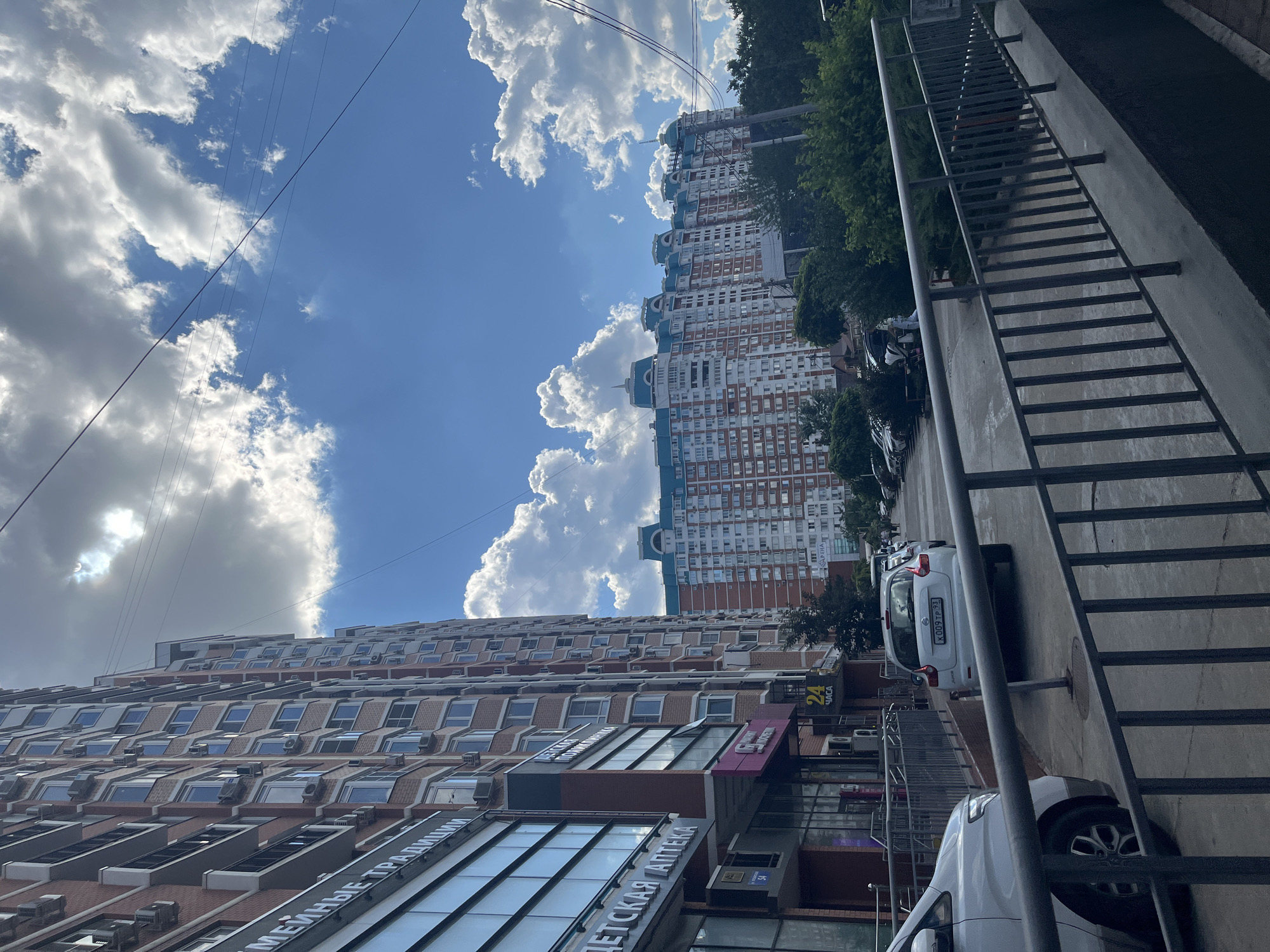 The image depicts a street view in Krasnodar, Russia, with a tall, brick apartment building on the left and another large, multi-story apartment building with a distinctive blue roof in the background. The sky is blue with fluffy white clouds. Power lines are visible crisscrossing the sky. In the foreground, there is a paved walkway with a metal railing. Several cars are parked along the street, including a white hatchback with a license plate visible. A few people are walking on the sidewalk. Businesses, including a "ДЕТСКАЯ АПТЕКА" (Children's Pharmacy) and another establishment with a "24 ЧАСА" (24 Hours) sign, are located on the ground floor of the building on the left. The overall scene suggests a residential urban environment.