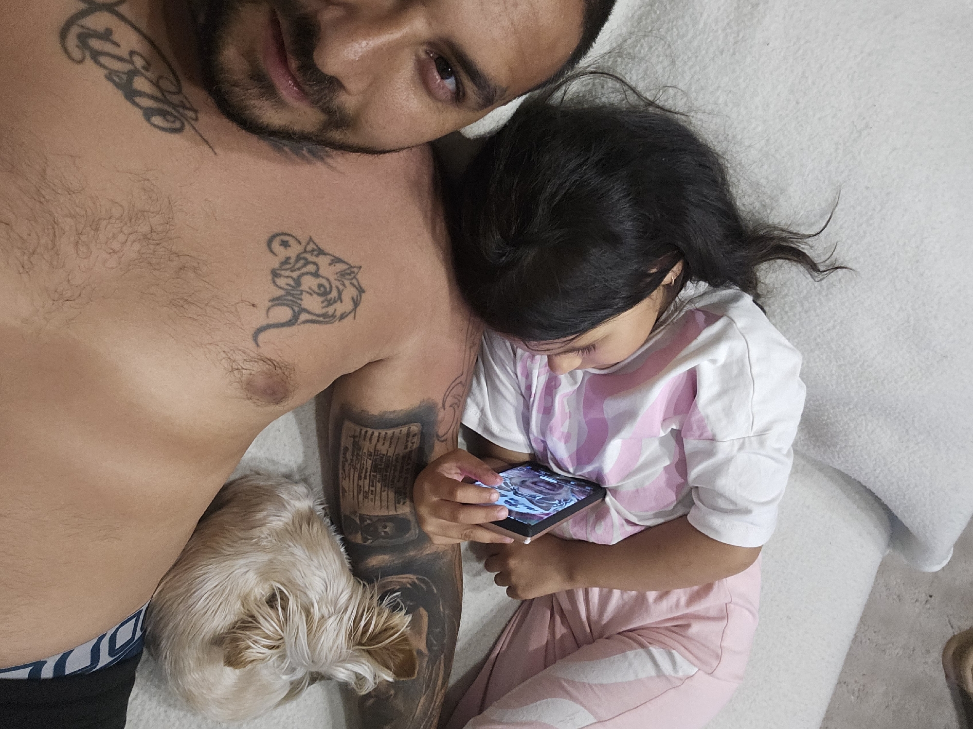 A low-angle, close-up shot captures a man and a child lying down on a textured white surface, possibly a sofa or bed. The man is positioned on the left, facing the camera, with his chest and face visible. He has dark hair, a beard, and several tattoos on his chest and arm. A small, light-colored dog is curled up on the man's chest, near his shoulder.
The child is lying on the man's chest, with their head resting on his shoulder. They are wearing a white t-shirt with pink stripes and pink and white striped pants. The child's hand is holding a smartphone, which is displaying an image. The child's eyes are closed, suggesting they are asleep or resting.
The overall environment appears to be an indoor setting. There is no information in the image to suggest a specific location within the city of Sofia, Bulgaria.