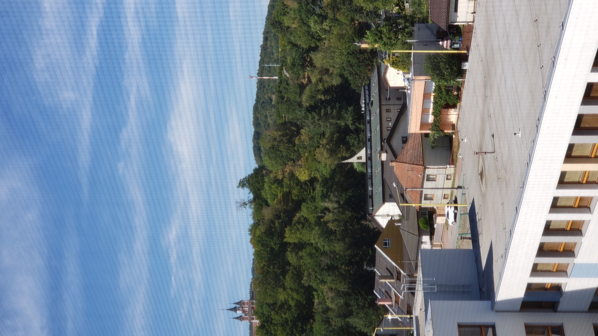 This is a vertical, medium-angle, daylight, outdoor photograph taken from an elevated position looking over a town. The sky is a bright blue with thin, white cirrus clouds. In the foreground, the flat, light-colored rooftop of a modern building with many rectangular windows runs across the bottom of the frame. Adjacent to this building, the pitched, red-tiled roofs of older, smaller residential buildings with various colored facades are visible. A white car is parked on a street running between these buildings, with yellow poles and streetlights lining the road. Behind the town, a lush, tree-covered hill rises, dotted with various buildings, including a large red-roofed building with two distinct spires at its peak, and a red and white radio tower further up the hill. The scene depicts a residential area with a backdrop of natural landscape, suggesting a town situated in a hilly region. The specific location is the area near the Kalvária church in Prešov, Slovakia.