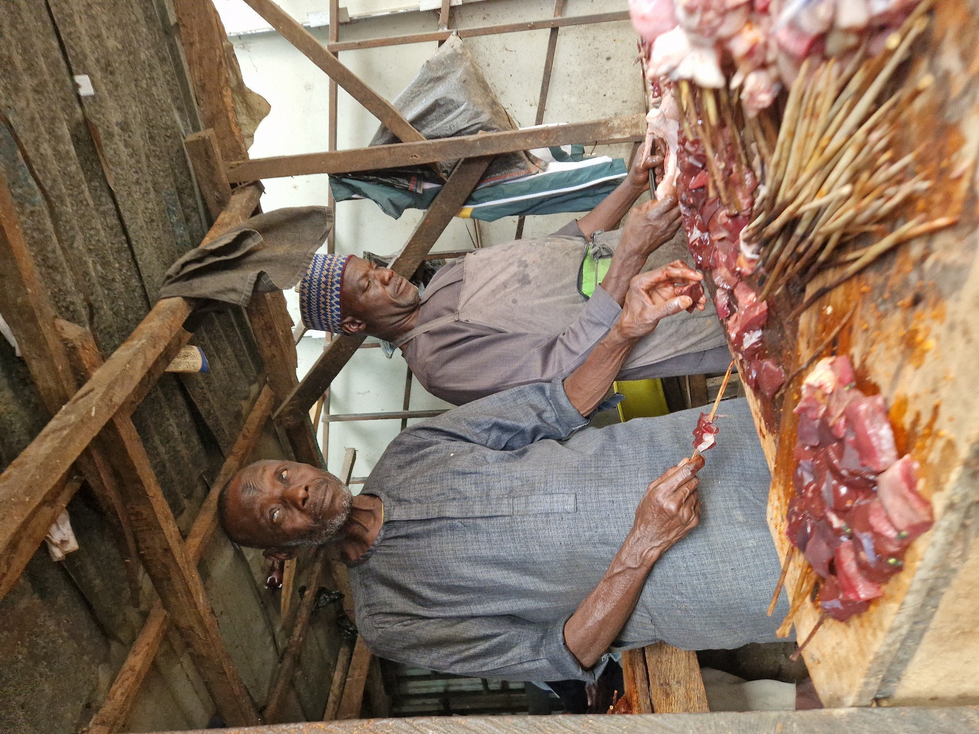 The media file depicts an outdoor market scene, likely in a warm climate given the attire of the subjects. Two men are the primary subjects. The man in the foreground, wearing a grey traditional garment, is holding a skewer with pieces of raw red meat, seemingly preparing it. His expression is neutral, with his gaze directed downwards. He has a beard and appears to be of African descent.
The second man, in the background, wears a purple and blue patterned cap and a light brown tunic. He is leaning and appears to be looking at the meat. His hands are also visible, suggesting he might be assisting or observing the meat preparation.
The scene is rustic, with wooden structures and corrugated metal roofing visible. A large wooden board is laden with more skewers of raw red meat. The meat is dark red, some pieces appear to be organ meat. The overall impression is of a butcher or meat vendor at a marketplace. The lighting suggests it is daytime, with no specific indicators of the weather. The focus is on the preparation of the meat, highlighting the raw ingredients and the hands-on process involved.