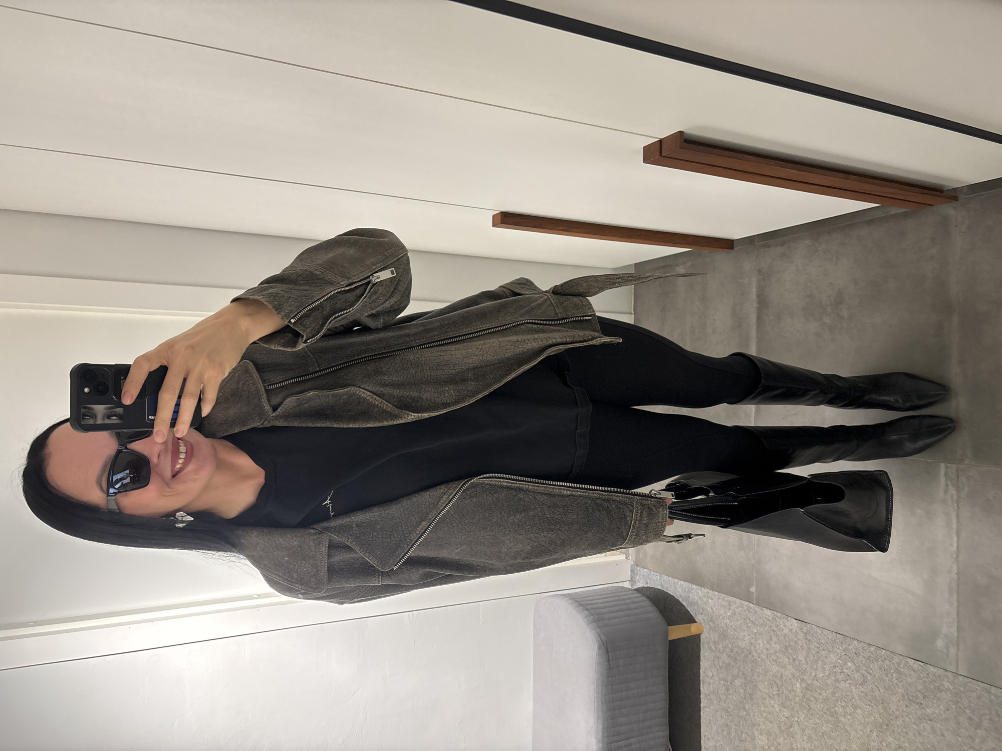 A woman is taking a selfie in a fitting room. She is wearing sunglasses, a black t-shirt, black pants, black boots, and a gray jacket. She is holding a black purse. The fitting room has white walls, gray flooring, and a gray bench. There are wooden beams on the ceiling. The location cannot be confirmed as Chaikovsky, Russia, based on the image alone.