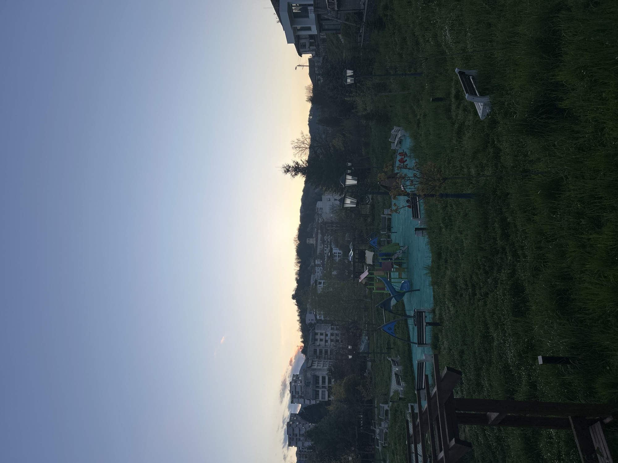 This landscape image, taken in Zlatograd, Bulgaria, depicts a quiet outdoor scene during either sunrise or sunset. The sky, occupying roughly the top half of the frame, transitions from a clear, medium blue on the left to a lighter, yellowish-orange hue near the horizon on the right, indicating the soft light of dusk or dawn.
The horizon line reveals distant hills or a forested area, dotted with numerous multi-story buildings that appear to be residential or commercial structures. In the middle ground, a public park or common area unfolds, featuring a vast expanse of green grass. Towards the right side of the image, a colorful children's playground is visible, featuring blue and green equipment. Several white or light-colored park benches are scattered across the grassy area and near the playground. Multiple streetlights with classic lantern-style tops are also present within the park.
In the immediate foreground, a dark wooden structure, possibly a fence or part of another bench, partially obstructs the bottom edge of the frame. The scene is devoid of people, conveying a sense of tranquility and stillness. No specific activity or event is depicted. There is no visible text in the image.
