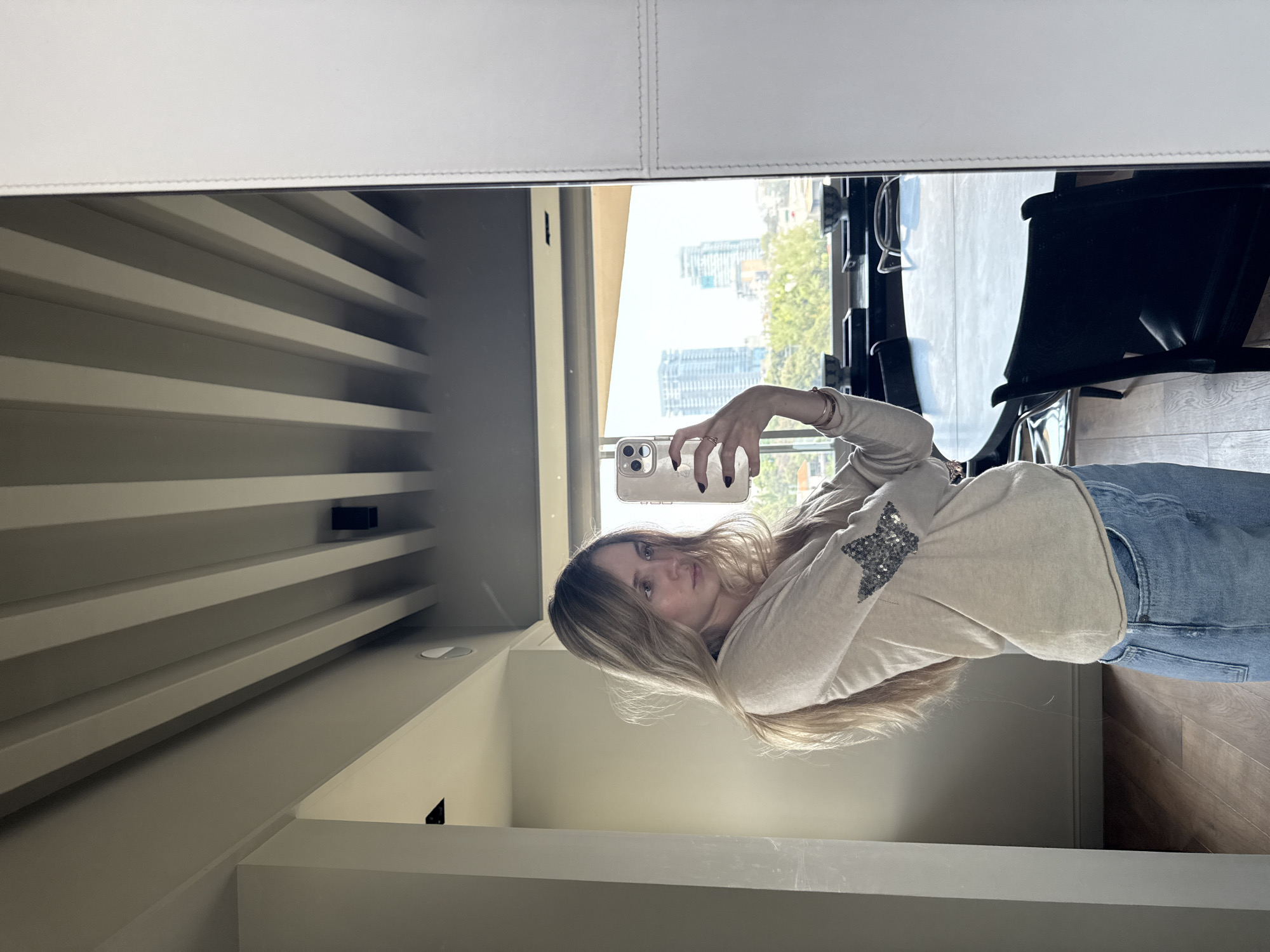 A woman stands in front of a large mirror, holding a smartphone, presumably to take a selfie. The reflection shows her blonde hair, a light-colored sweater with a star embellishment on the sleeve, and blue jeans. The mirror's reflection also includes a cityscape with tall buildings, suggesting an urban environment. In the room, a black chair and a white table are visible. The ceiling has a modern design with parallel lines. The image doesn't provide definitive location information to confirm it is Mexico City, Mexico.