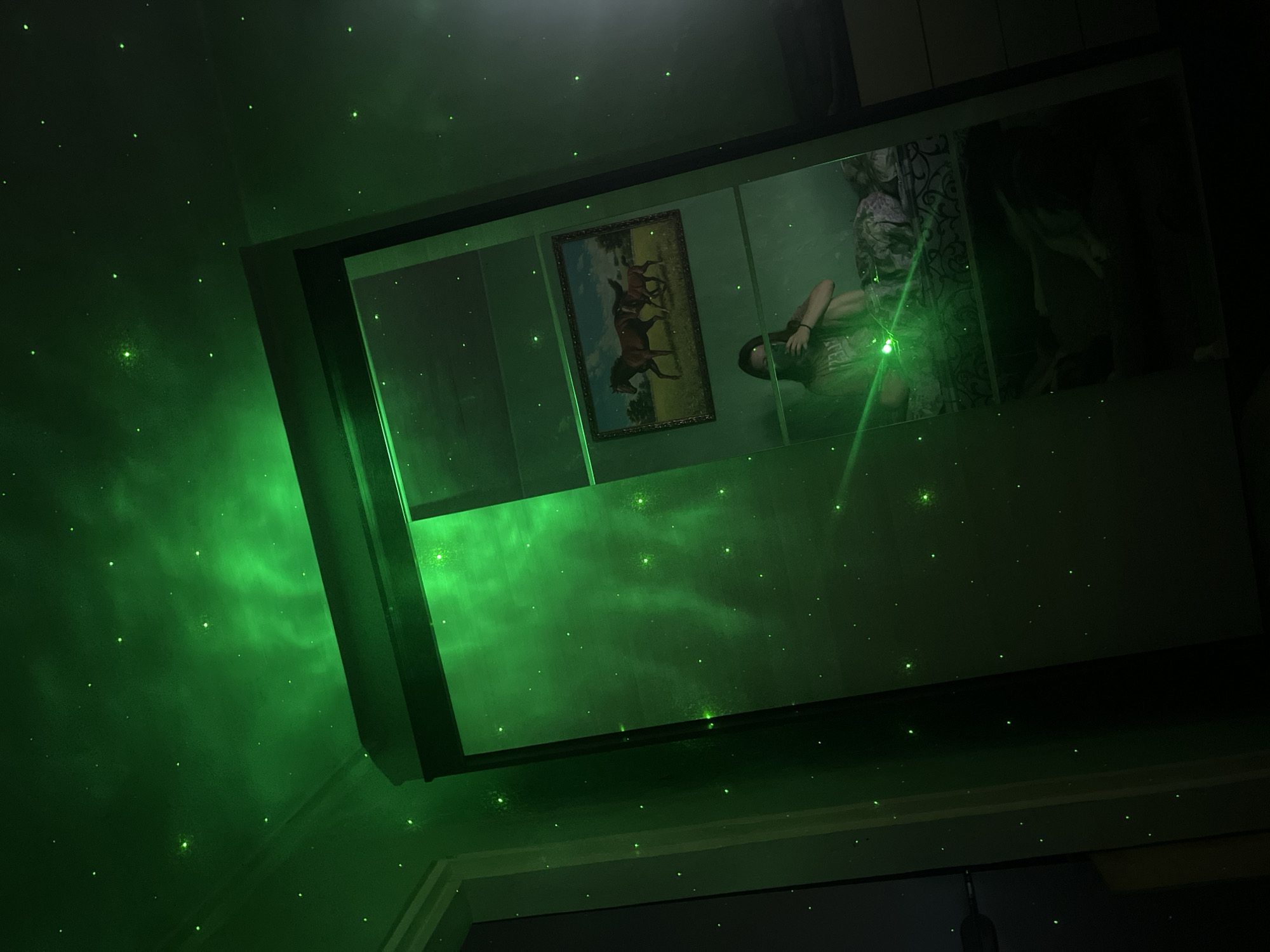 A room is illuminated with green star-like lights projected onto the walls and ceiling. A rectangular mirror reflects the scene, including a painting of horses on the wall, a person lying down with a phone pointed upward, and curtains behind the person. The room appears to be in a residential building. There is no mention of a city or location, and therefore no determination that the location is Nizhny Tagil, Russia.