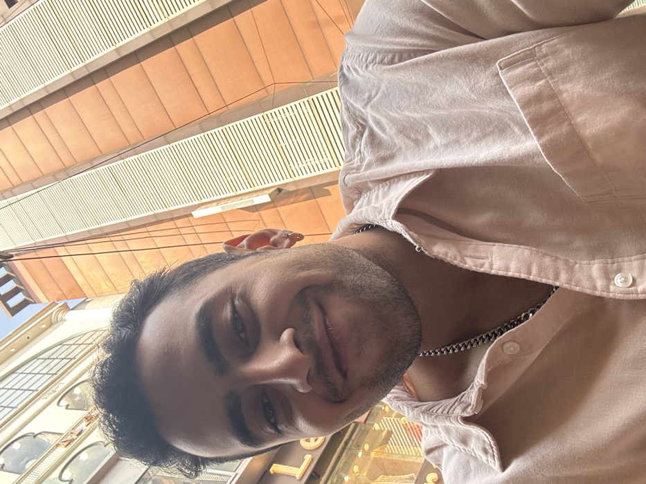 A man is captured in a close-up selfie, tilting his head slightly and smiling with a relaxed expression. He has short dark hair and a neatly trimmed beard or stubble. He is wearing a light-colored, possibly salmon or beige, button-down shirt, unbuttoned at the top to reveal a silver chain around his neck. A small, reddish, ear-shaped device, possibly a hearing aid or an in-ear monitor, rests on top of his head near his ear.
The photo is taken outdoors during bright daylight, indicated by the clear lighting and visible shadows. The background features urban architecture. Directly behind and above the man are modern buildings with warm, possibly orange or terracotta-toned, facades and white horizontal slats or railings. To the lower left, an older building with ornate details and arched windows is visible, suggesting a blend of architectural styles. The overall perspective is tilted, creating a dynamic diagonal composition. The scene suggests a bustling city environment, consistent with Kolkata, India. Partially visible, indistinct letters like "LE" from a sign can be seen in the bottom right corner.
