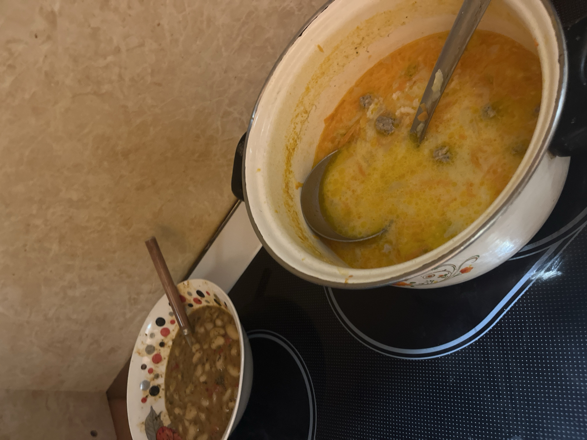The image depicts a kitchen scene, possibly in a domestic setting. A pot filled with soup, presumably a yellow-colored broth with what appear to be meatballs, sits on a modern black stovetop. A metal ladle rests inside the pot. To the left, a smaller bowl containing a bean-based dish is visible, along with a spoon. The countertop behind the stovetop has a light brown, mottled pattern. The exact location in Burgas, Bulgaria cannot be determined from the image. No people are directly visible.