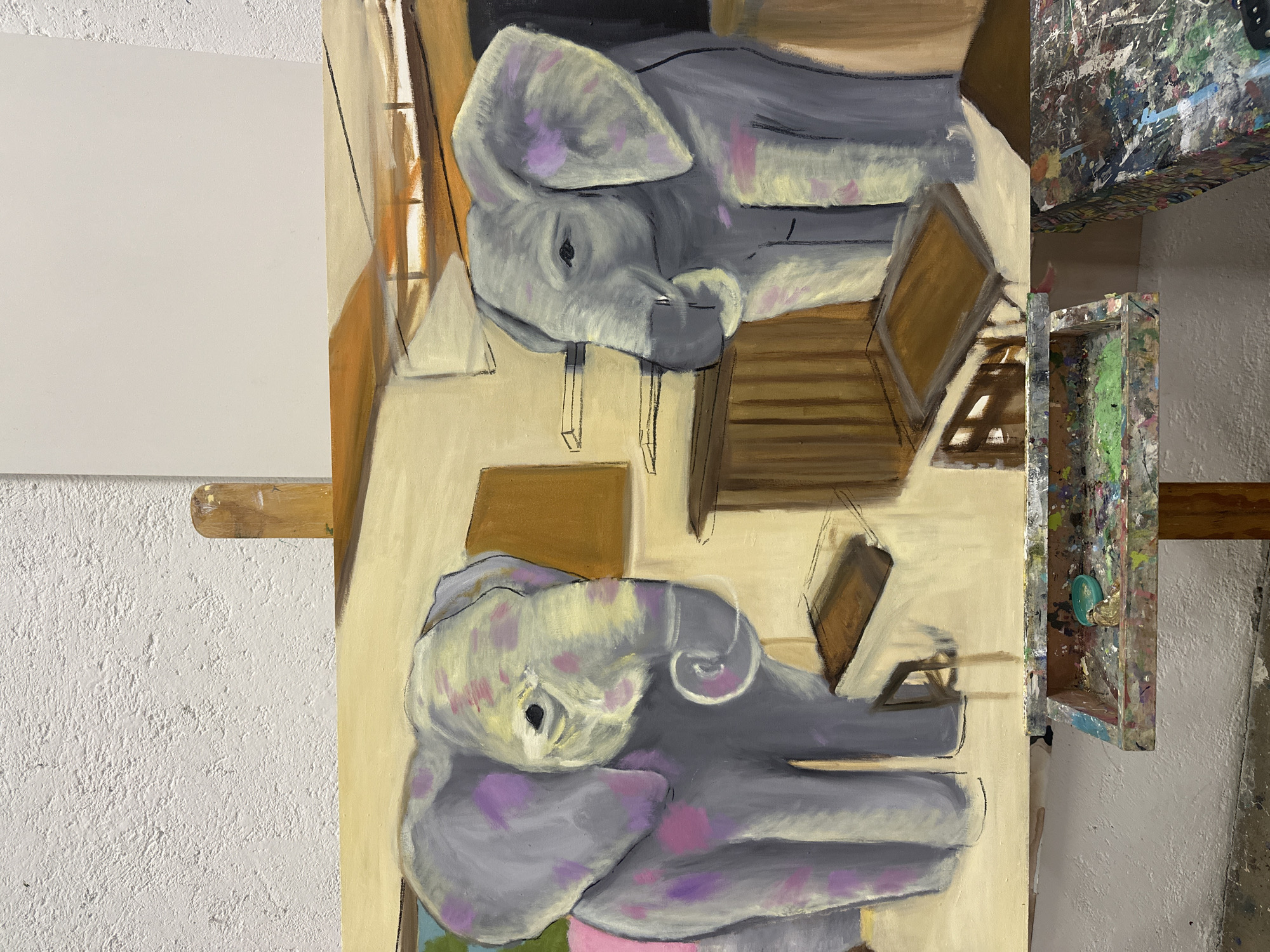 The image shows an unfinished painting of two grey elephants in an indoor setting. The elephants are stylized with pink and yellow splotches and appear to be positioned one above the other, almost mirroring each other. They are surrounded by what appear to be simplified depictions of furniture or architectural elements in shades of brown and beige. The painting is on an easel. To the left of the painting is a blank white canvas also on an easel. To the right of the painting, and slightly in the foreground, are painting accessories, including what appears to be a tray or palette. The background consists of a white wall. The location in the city of San Bernardino Tlaxcalancingo, Mexico is not able to be confirmed from just this image.