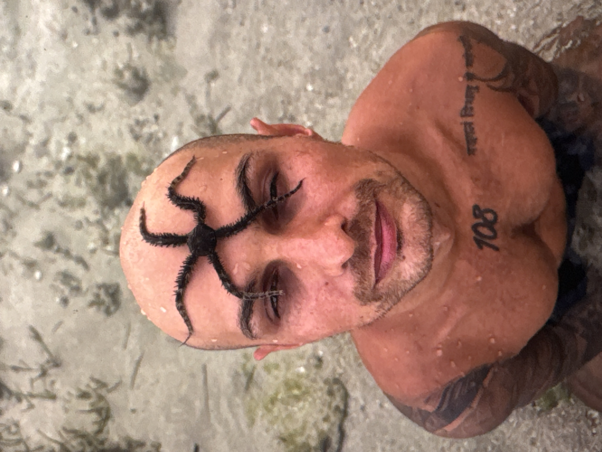 A man with a shaved head and a goatee is in shallow water, with a brittle star on his head. He is looking up at the camera. The man has tattoos on his arms and chest, including the number "108" and some text in an Indian script on his chest. The water is sandy with some aquatic plants. The image was taken in Kuta Selatan, Indonesia.