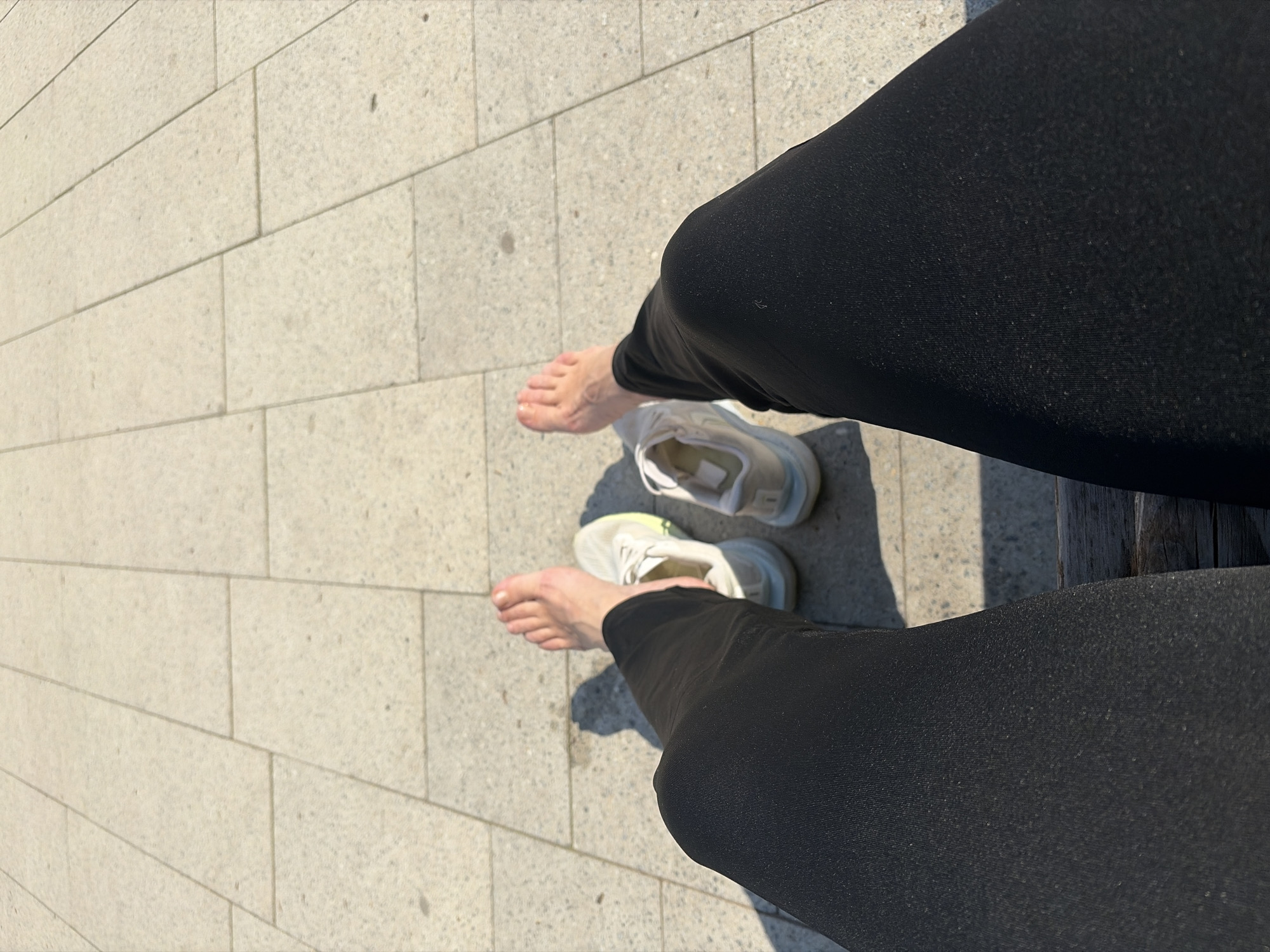 The image shows a person sitting on a bench with their shoes off in an outdoor setting, likely a public space paved with rectangular stones. The person is wearing black leggings. Their bare feet are visible next to a pair of white sneakers. The location is purportedly Chaikovsky, Russia, though this cannot be confirmed from the image alone. The scene is sunlit, casting shadows on the pavement. The angle of the photo is from above, looking down at the person's legs and feet.