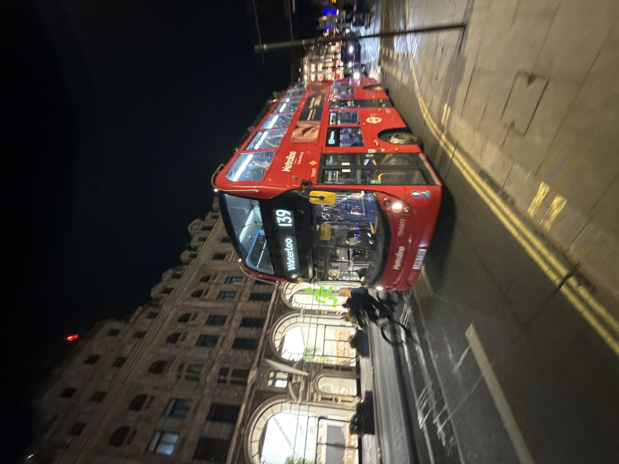 At night in London, a red double-decker bus, route 139 to Waterloo, is seen traveling along a city street. The bus's destination is displayed prominently on its front. A cyclist rides nearby, possibly in the same direction. The street has double yellow lines indicating parking restrictions. Buildings with illuminated arched windows line the side of the street, with a taller building in the background. The lighting suggests it is nighttime.