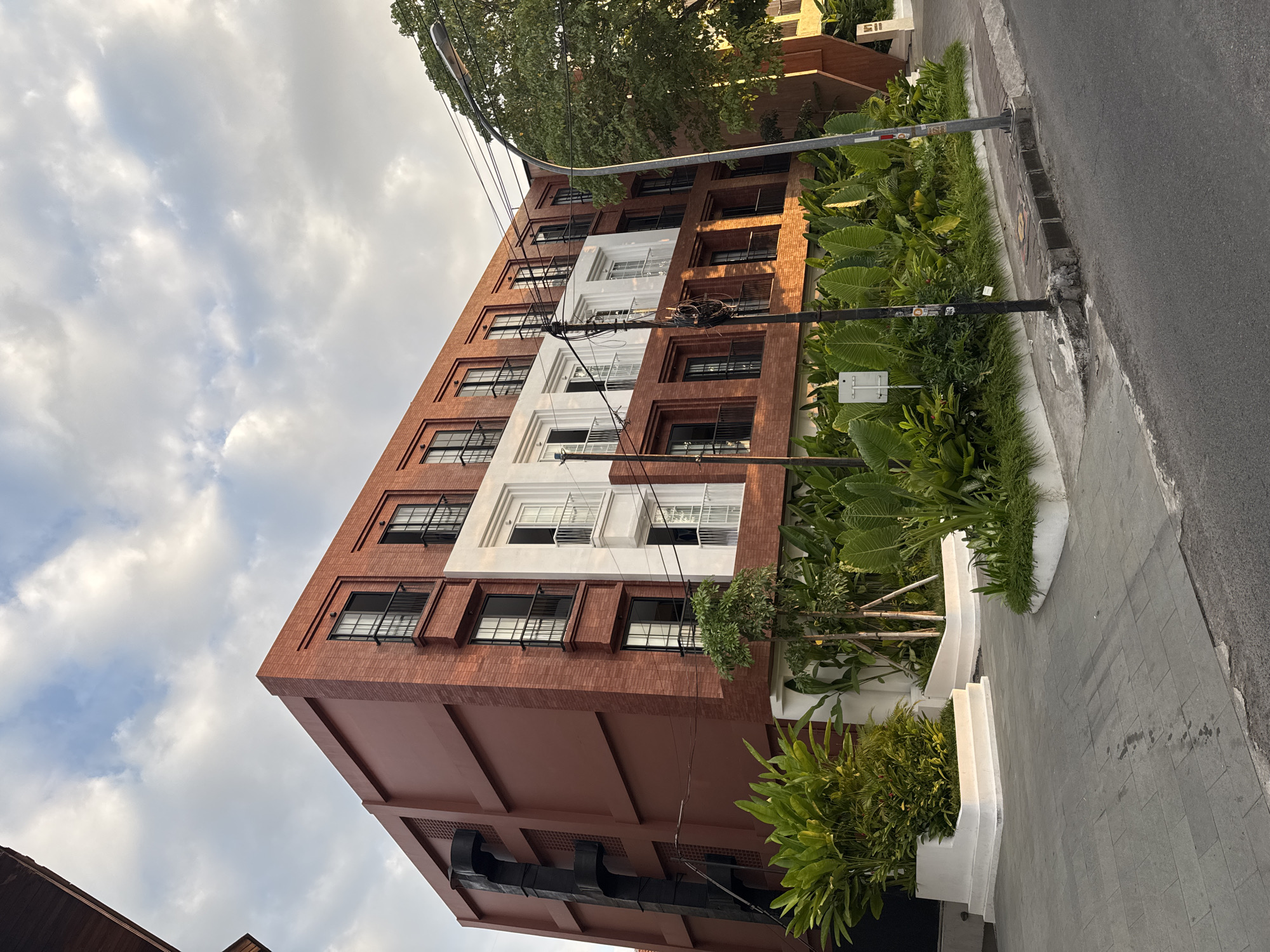 In Kuta, Indonesia, a multi-story modern building with a red brick facade and white accents stands along a paved street. The building features numerous balconies with black railings and windows. Lush green plants are arranged in white planters in front of the building and along the sidewalk. A street lamp and electrical wires are visible on the right side of the image. The sky is partly cloudy, with the sun casting a warm light on the scene.