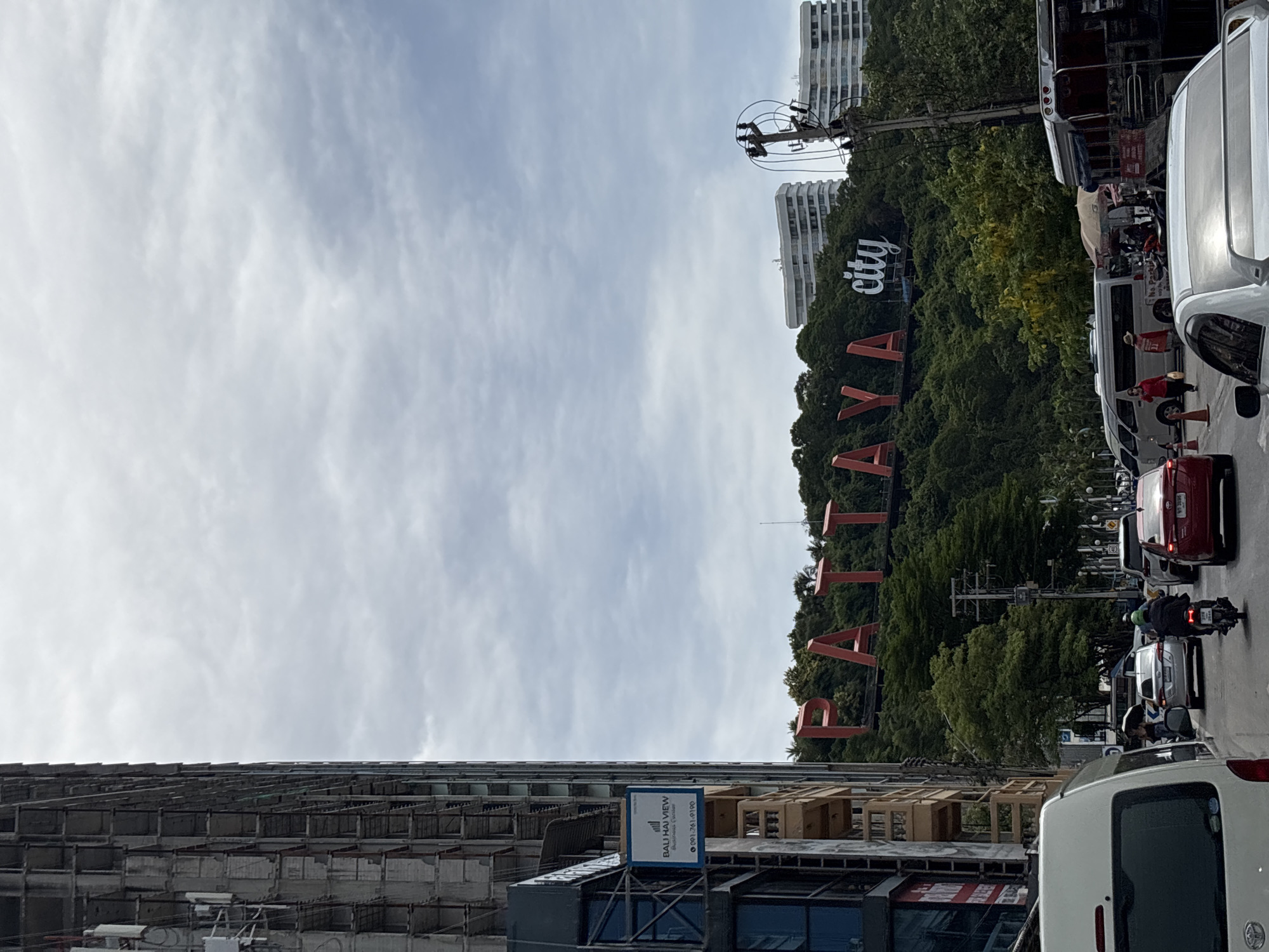 The image shows a street scene in Pattaya City, Thailand, with a prominent "PATTAYA city" sign on a hill in the background. To the left, an unfinished or under-construction building with exposed concrete and scaffolding dominates the foreground. A blue sign on this building reads "BALI HAI VIEW" and a phone number. The street is busy with traffic, including cars, motorcycles, vans, and a bus. Several people are visible on the street and sidewalk, some walking, some riding motorcycles. The sky is overcast with clouds. The overall impression is of a bustling urban environment with ongoing development and a famous landmark.
