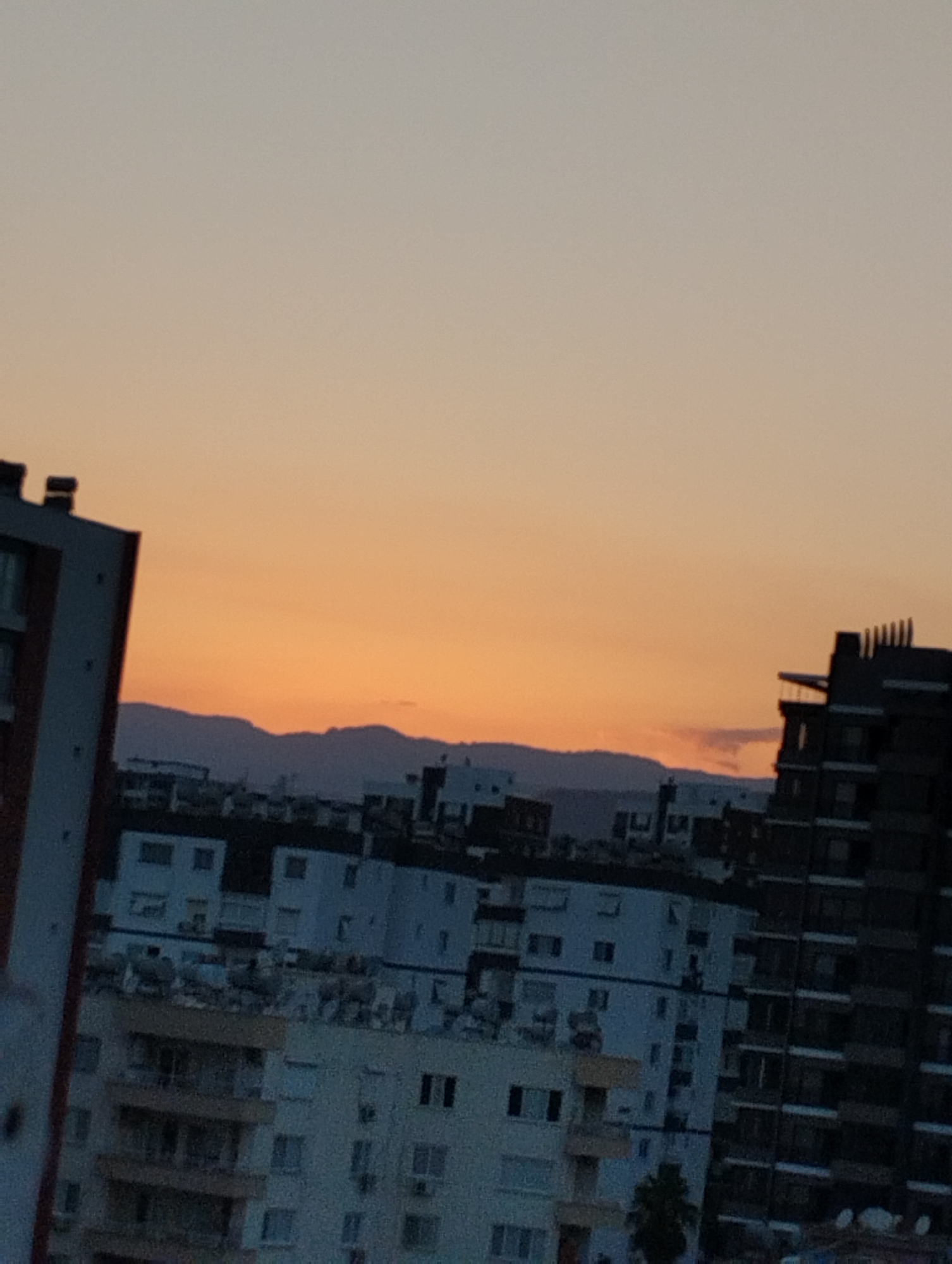The image depicts a city skyline at sunset, likely in Mezitli, Turkey, characterized by a gradient sky transitioning from a pale orange near the horizon to a muted gray at the top. Silhouetted against the sky are several multi-story buildings of varying architectural styles and heights. On the left, a dark building with red trim is partially visible. In the center, a collection of lighter-colored residential buildings with numerous windows are clustered together. To the right, a taller, dark building with a distinctive, stepped facade and rows of windows dominates the scene. In the distance, a range of low, dark mountains is visible, their outlines softened by the atmospheric haze. The overall impression is one of an urban environment at dusk, with the natural beauty of the sunset contrasting with the man-made structures.