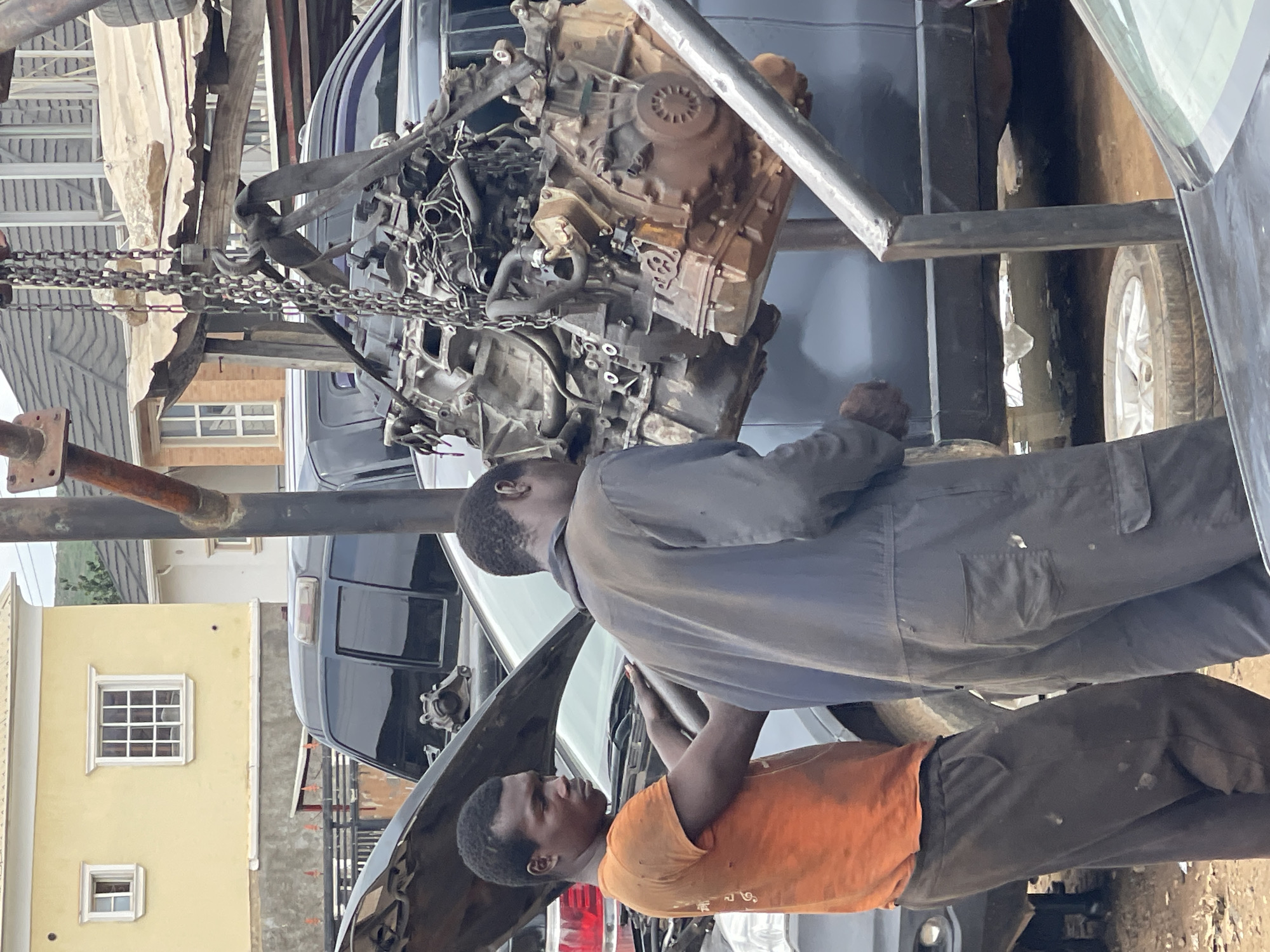 The image depicts an outdoor auto repair scene in Abuja, Nigeria. Two men, appearing to be mechanics, are engaged in the process of removing or installing a car engine. The engine, a complex assembly of metal parts and wiring, is suspended by a chain hoist and positioned between the two individuals. One mechanic, wearing an orange t-shirt and dark pants, is positioned to the left, looking towards the engine and appears to be steadying it with his hands. The second mechanic, dressed in dark blue overalls, is on the right, facing away from the viewer and also near the engine. They are working in an area with several vehicles present, including a dark SUV with its hood open, and another vehicle partially visible in the foreground. The environment suggests a busy workshop, with dirt and debris on the ground, and a rustic overhead structure providing some shade. The background shows residential buildings with distinct architectural features typical of Abuja. The overall atmosphere is one of practical, hands-on labor within a functional urban setting.