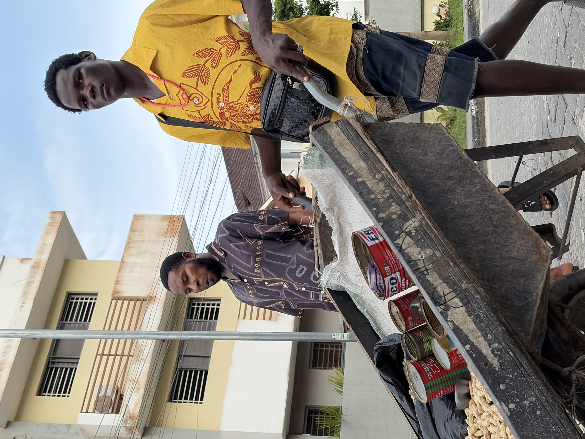 In Igboefon, Nigeria, a person pushes a wheelbarrow filled with peanuts and several cans. The wheelbarrow is old and shows signs of wear, with dirt and scratches on its surface. The contents include a pile of peanuts in shells, along with an assortment of tin cans, some of which are open. One can is labeled "TOMATO" with a red and green design, another has red and yellow branding that appears to read "PE PASTE". A third can has a red and white label with the word "ER" prominently displayed. The person pushing the wheelbarrow is wearing dark shorts with a patterned trim and a bright yellow short-sleeved shirt. They are also wearing a red beaded necklace and a black crossbody bag. Another man, standing to the left of the wheelbarrow, is wearing a patterned dark shirt. In the background, a multi-story building with balconies and windows is visible, along with utility poles and wires, suggesting an urban setting. The sky is overcast with a pale blue hue.