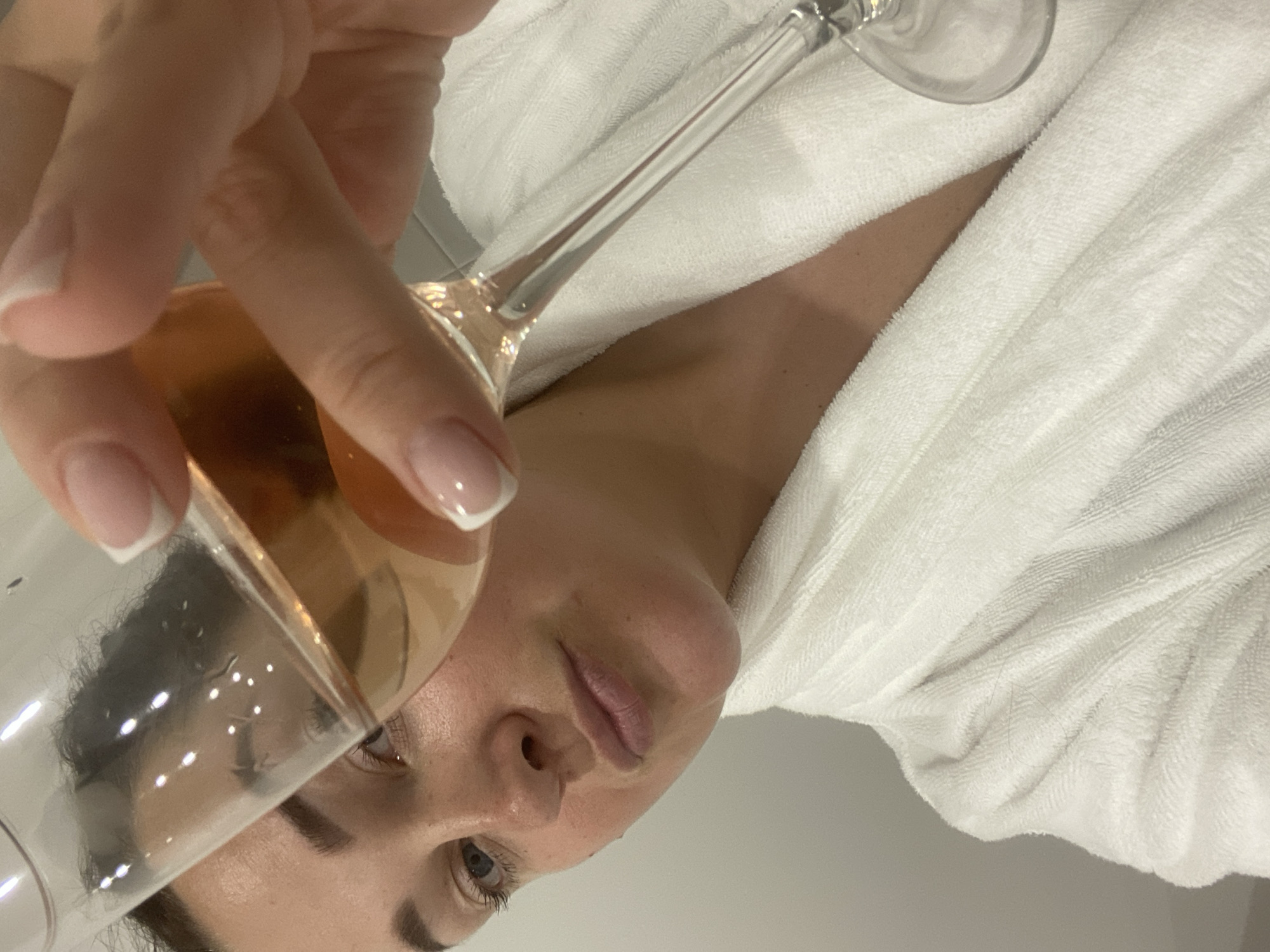 A woman is holding a glass of rose wine close to her face, and she appears to be wearing a white terry cloth robe. The background is a plain white wall. There are no other visible subjects or objects. The location is not discernible from the image.