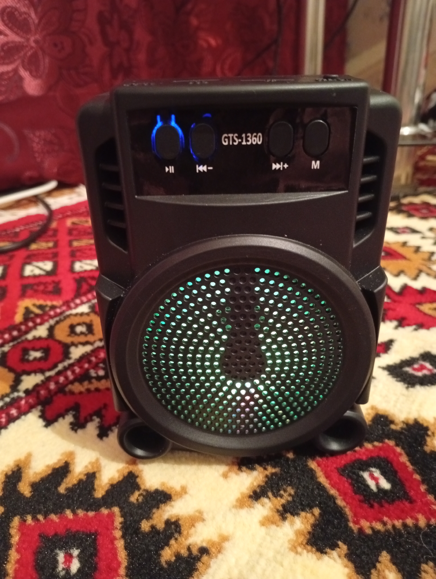 A black portable speaker with a lit green speaker grille is placed on a red and beige patterned carpet. The speaker has several control buttons on top, including a power button, volume controls, and playback buttons. A red fabric with a floral pattern is visible in the background. The image is a close-up shot of the speaker, with the surrounding environment out of focus. There are no people or animals in the image. This scene is not specific to Seýdi, Turkmenistan, or any particular city.