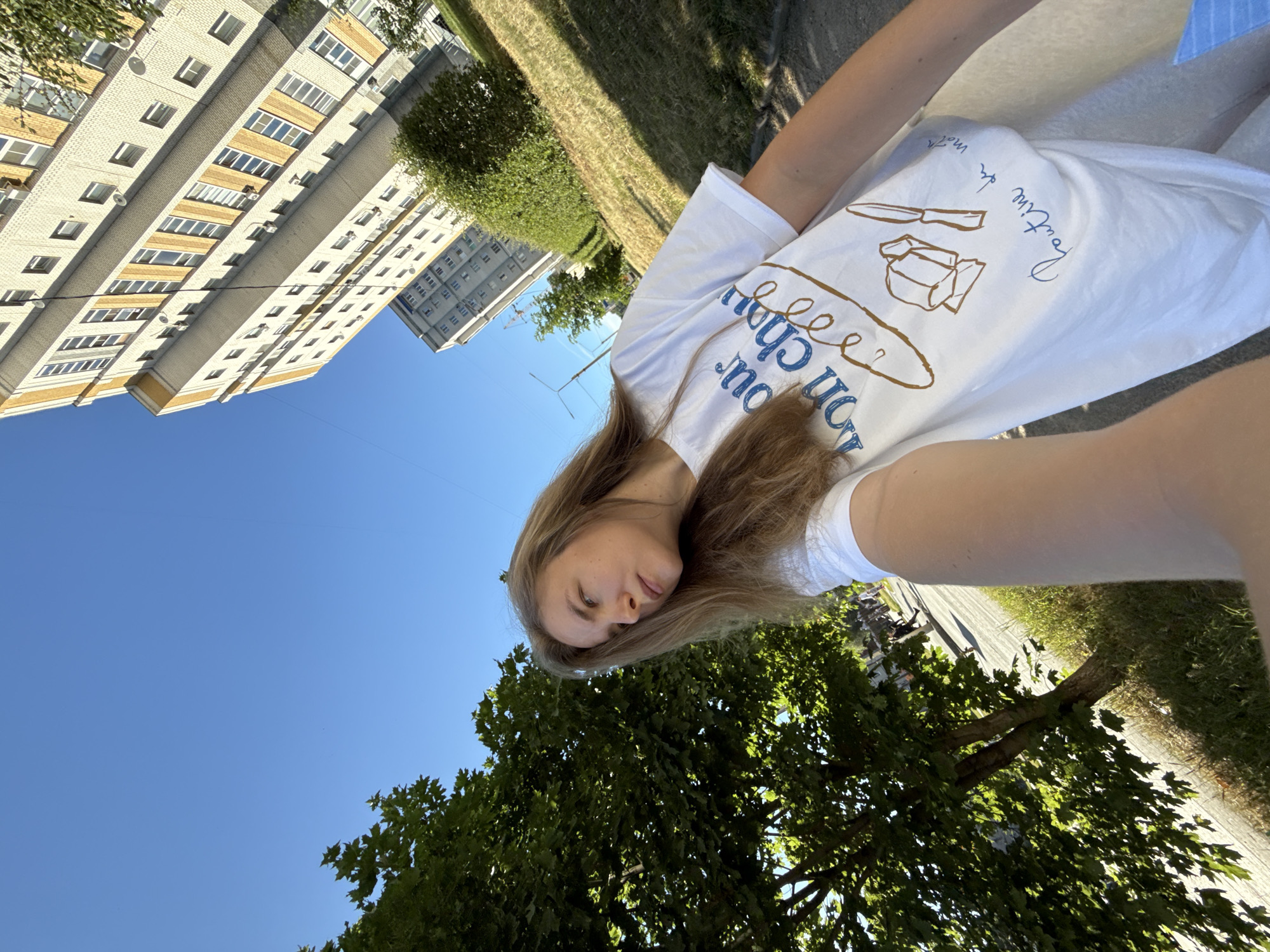 In a sunlit, outdoor urban environment, a young woman with long, light brown hair, wearing a white t-shirt with blue and brown graphics, takes a selfie. She is positioned in the foreground, with her face turned to her right, looking downwards. Behind her, a large green tree with lush leaves dominates the left side of the frame. In the background, a tall, multi-story residential building with a light-colored brick facade and yellow accents is visible against a clear blue sky. To the right of the tree and in front of the building, a grassy slope runs alongside a paved walkway, where a few indistinct figures can be seen. The lighting suggests it is daytime, likely late afternoon, given the long shadows. The specific location is identified as the city of Vladimir, Russia.