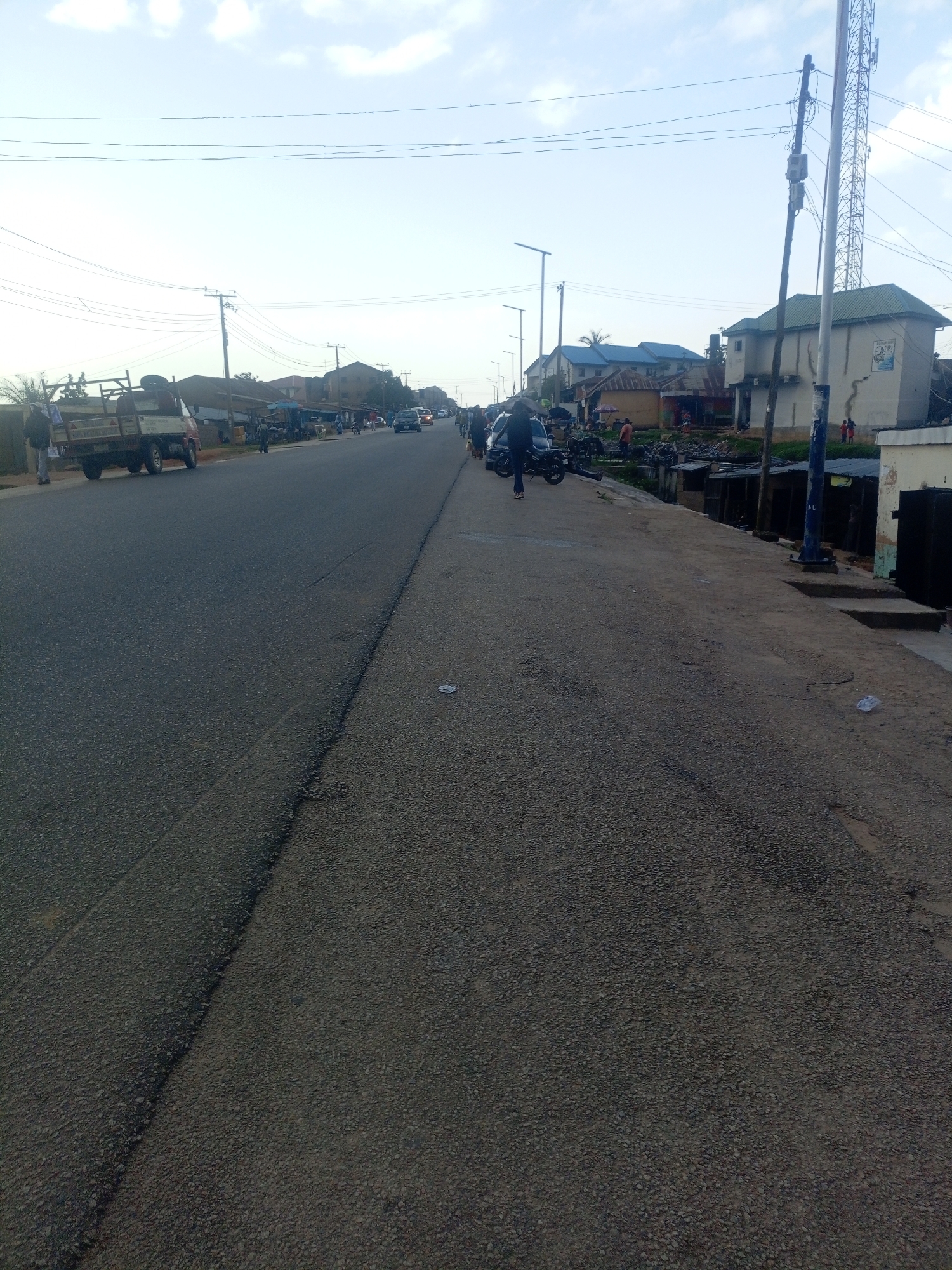 The image shows a street scene in Jos, Nigeria, with a paved road running horizontally across the frame. To the left of the road, there is a white flatbed truck, and further down the road, a group of people are walking. On the right side of the road, there are buildings, including a two-story structure with a green roof and a telecommunications tower. The sky is cloudy. The scene appears to be a busy commercial area, with several businesses and stalls lining the street.