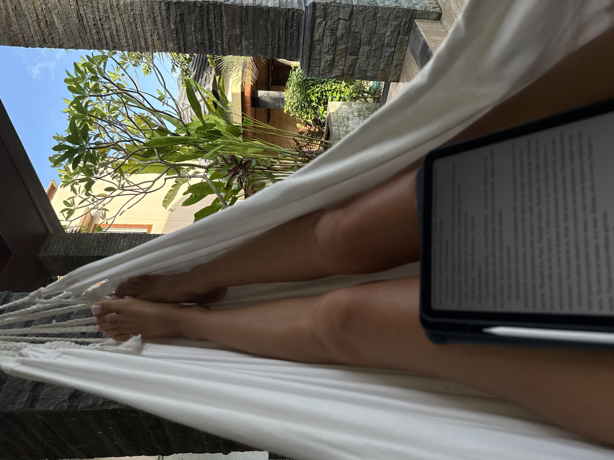 The image depicts a person lying in a white hammock outdoors in Kuta Selatan, Indonesia. The individual is positioned between two stone pillars. The person's legs are visible extending out along the hammock's length, with their feet at the left side of the image. A tablet, with text visible on the screen, rests on the person's legs. A white stylus is visible next to the tablet. In the background, beyond the hammock, a tropical garden with trees and plants is visible. In the distance, a building with a yellow exterior is observed. The sky is partly visible through the trees.