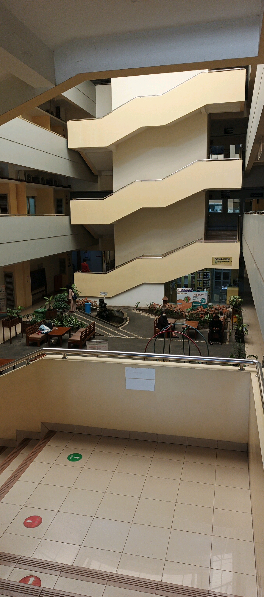 The image shows the interior of a multi-story building with a central open atrium featuring a prominent, cream-colored staircase that zigzags upwards. The building's architecture is characterized by exposed concrete and painted walls in neutral tones, with multiple balconies and walkways overlooking the atrium.
On the ground level of the atrium, there is a small landscaped area with potted plants, wooden benches, and tables. A dark, sculptural fountain is visible in this area. Two individuals are seated on benches.
On the floor of the atrium, in the foreground, there are light-colored tiles with a grid pattern. Several circular floor markers are visible, including a green one with an arrow pointing forward, and red ones with arrows. Steps are also visible leading down to this tiled area.
In the background, on the upper levels, windows and doors are visible, suggesting office or room spaces. A sign with "POJOK ALKES" is affixed to one of the walls near the staircase. The overall impression is of a functional, possibly institutional, building with an emphasis on natural light and open space. The presence of the floor markers suggests a current operational protocol, potentially related to crowd management or directional guidance. The location is identified as Ambarketawang, Indonesia.