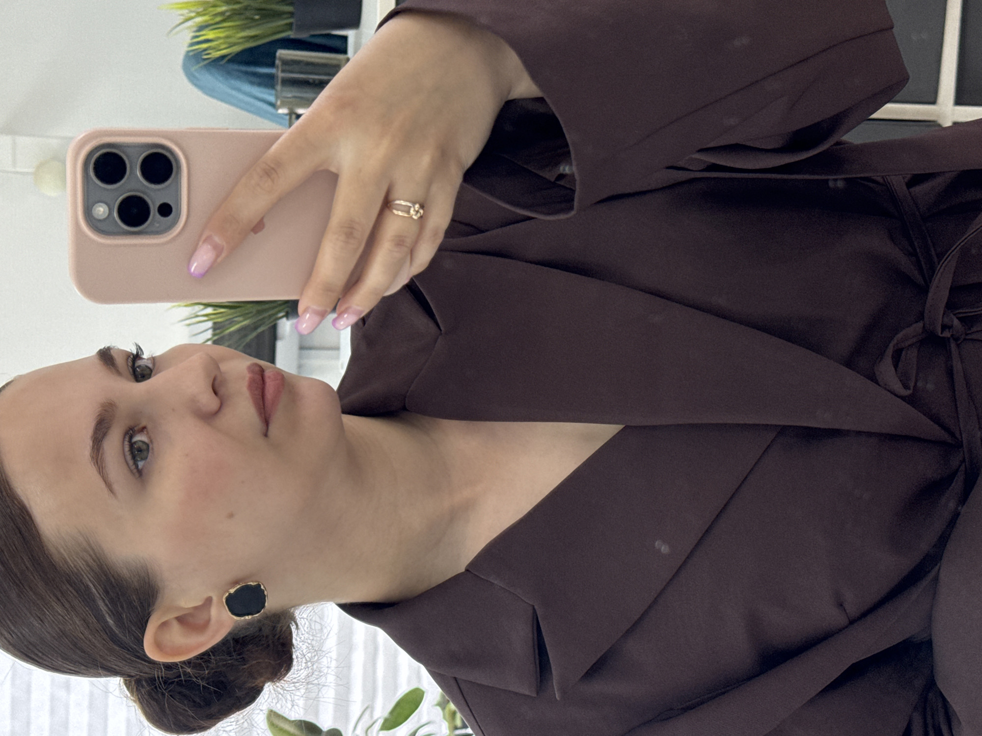 A woman is taking a selfie in a mirror. She is wearing a dark brown blazer and has dark hair pulled back into a bun. She has on black earrings and a gold ring on her right hand. Her fingernails are painted a light pink. In the background, there are plants and what appears to be a blue cloth. The image does not contain any identifiable landmarks or elements that would suggest its location in Chaikovsky, Russia.