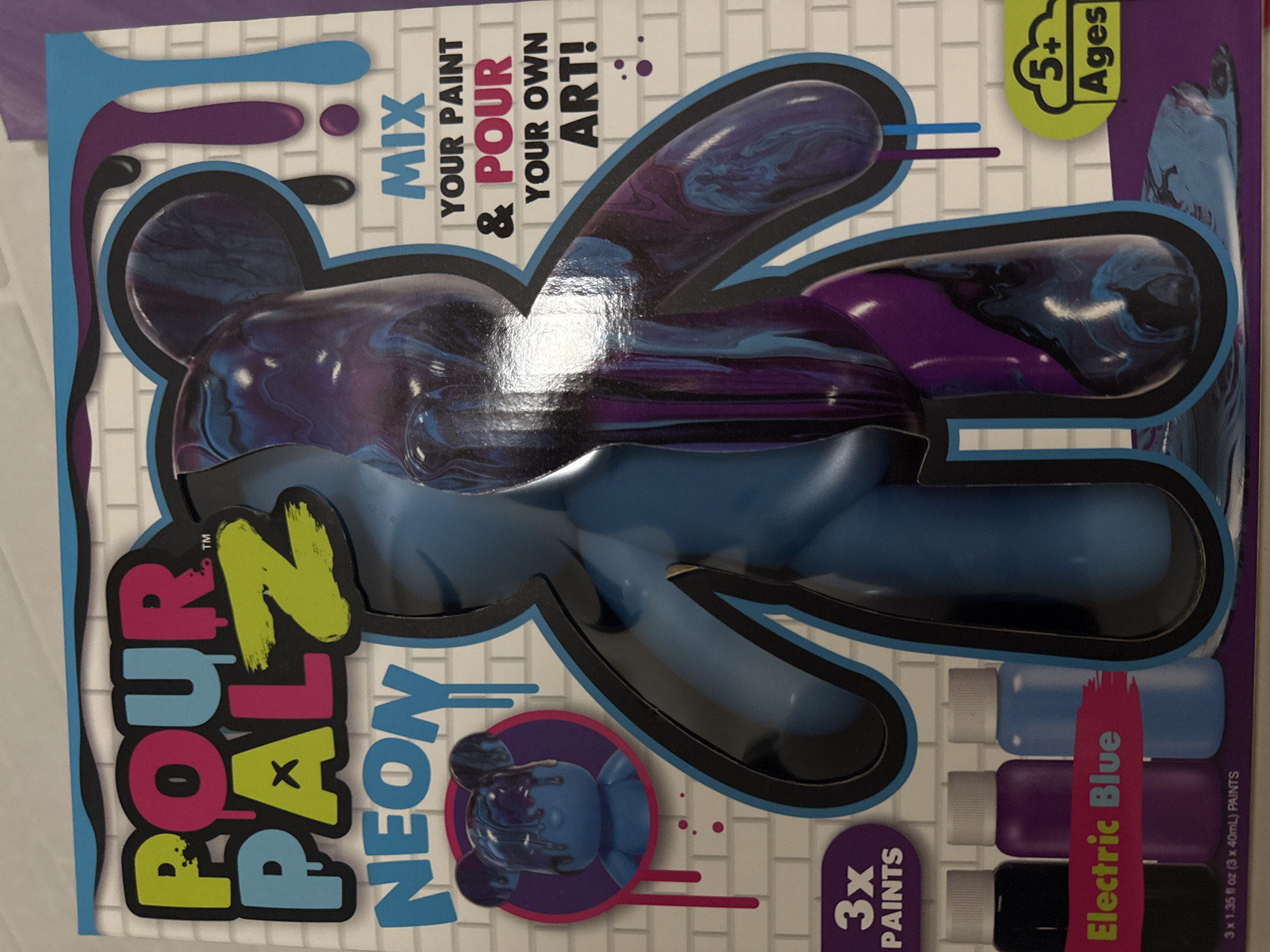 This image displays the packaging for a children's craft kit called "POUR PALZ NEON." The main subject is a product box featuring a large, transparent window that showcases a stylized, bear-like figure. One arm and part of the head of the figure inside the packaging are shown covered in swirled patterns of dark purple and blue paint, while the rest of the figure is unpainted in a solid blue.
The activity promoted is paint pouring, as indicated by the text "MIX YOUR PAINT & POUR YOUR OWN ART!" The kit includes three small bottles of paint visible on the lower right: light blue, purple, and black. A highlighted text strip next to the paint bottles reads "Electric Blue," suggesting one of the included colors.
Notable details include the bright, playful design of the packaging. The background features a subtle white brick pattern. Drip-like shapes in blue, purple, and black accent the top left corner of the box. A small circular image on the lower left provides an example of the finished product, showing the blue bear figure with poured paint on its head. The box clearly states "5+ Ages" in a green bubble, indicating the recommended age group. "3X PAINTS" is written, and below the paint bottles, "3 x 1.35 fl oz (3 x 40mL) PAINTS" specifies the quantity and volume. The product name "POUR PALZ NEON" is prominently displayed on the left side of the box in large, colorful letters (pink, blue, and lime green).