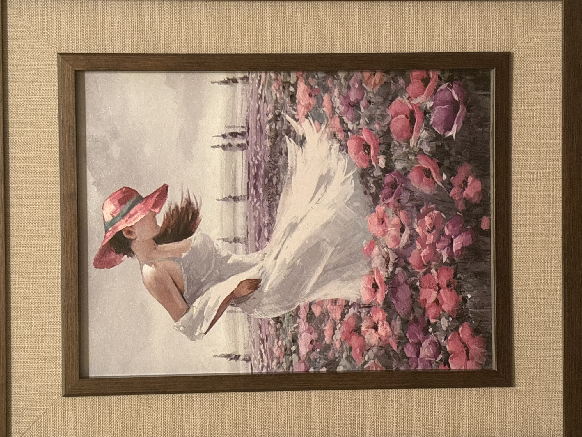 The image shows a framed painting. The painting depicts a woman wearing a pink hat and a white dress, seemingly caught in motion. The woman is positioned against a backdrop of lavender fields and a vibrant cluster of pink and purple flowers. The frame around the painting is dark brown, and it is set against a light beige textured background. The location is not specified and is not evident from the image. The painting is the subject of the image, and there is no information about the city of Montana, Bulgaria.