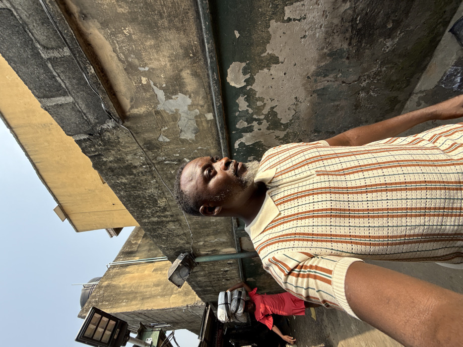 The image portrays an outdoor urban environment within the city of Ado, Nigeria, under clear, bright daylight. In the foreground and midground, an adult male with a salt-and-pepper beard and short dark hair is visible, oriented towards the viewer's left. He is wearing a short-sleeved, collared polo shirt featuring vertical stripes in cream, brown, and light blue/green, with a quarter-zip neckline. His right arm is partially visible, extending towards the lower left of the frame.
Behind the male, to the left, a younger individual in a red dress with ruffled sleeves is walking away from the camera, carrying a stack of white, bundled items on their head. Adjacent to this individual, a structure with visible wiring and a black floodlight fixture is present; a partial sign reading "WELCOME" is discernable.
The background is dominated by a weathered concrete block wall running parallel to the scene, displaying significant peeling paint or plaster and discolored patches of grey, green, and brown. Electrical wiring runs horizontally along the upper portion of this wall, and a smaller, grey floodlight is mounted on it. Above the concrete wall, a building with a yellow or ochre-colored facade is visible against a clear blue sky.