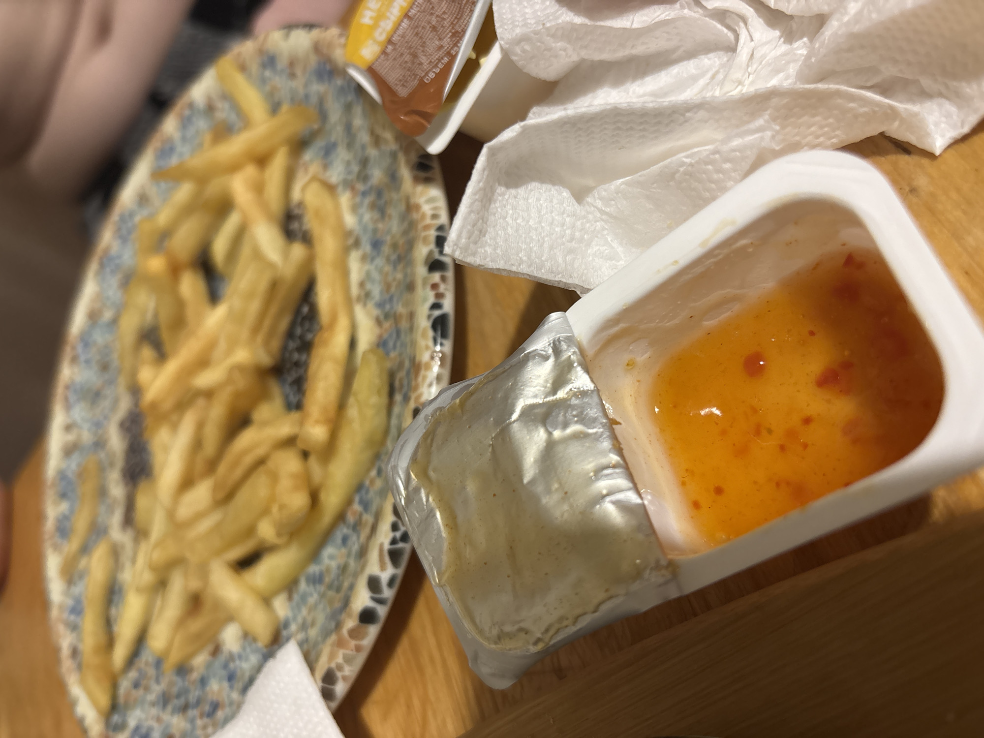 The image presents a casual dining scene on a wooden surface. A plate filled with french fries occupies the left side of the frame, its patterned design featuring blue and brown hues. Adjacent to the plate are several condiments: a small container holding a yellow sauce, a white condiment container with its foil lid partially peeled back, revealing a translucent orange-colored sauce with red specks, and a brown sauce packet labeled "HEP". White napkins are scattered between the plate and condiment containers. The scene appears to be set indoors, possibly in a home or restaurant. The city and country of the location are not known.