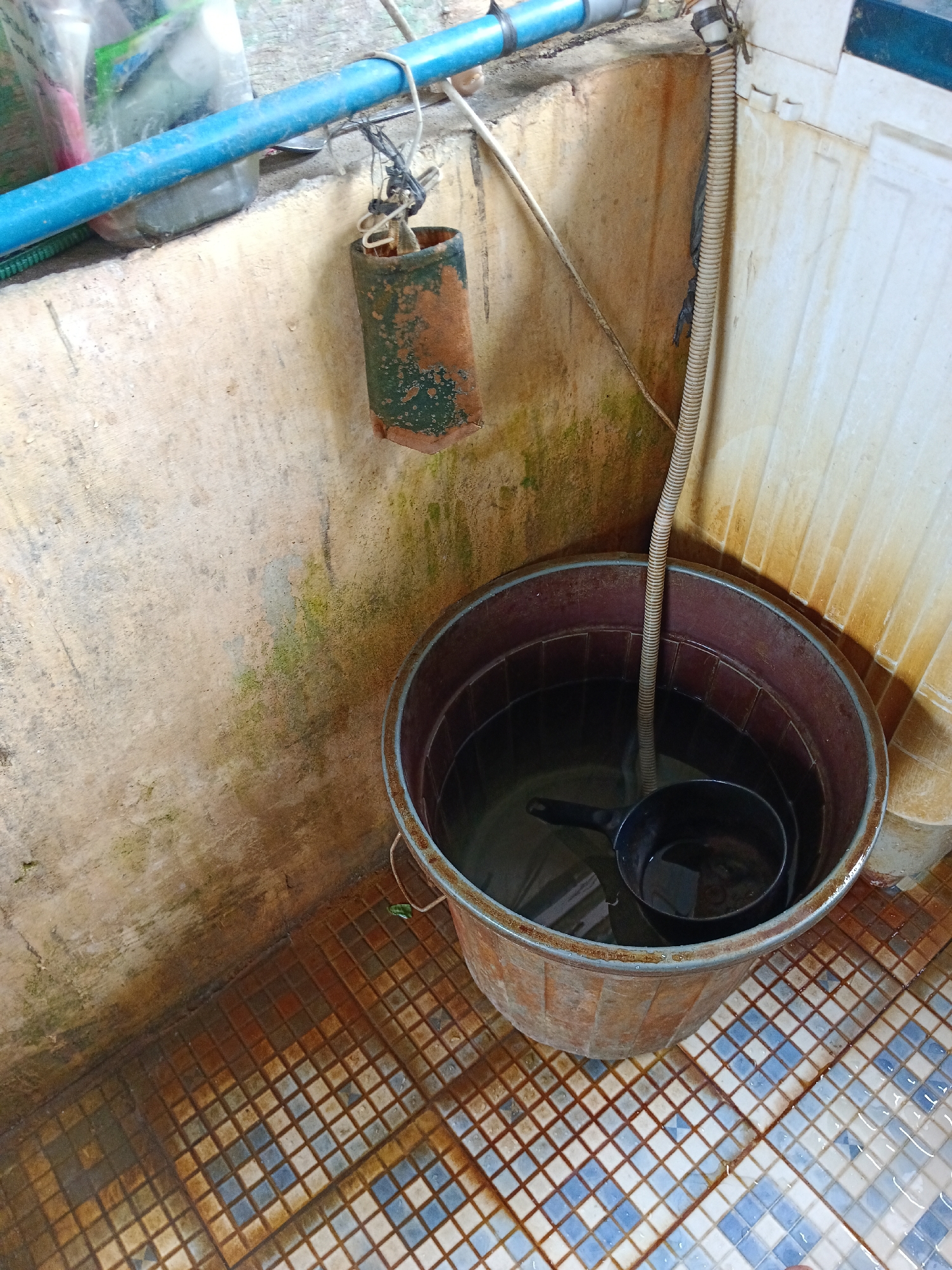 The image depicts an outdoor scene, likely a utility area or bathroom, in Aek Kanopan, Indonesia. A large, weathered, reddish-brown plastic tub is filled with water, holding a black plastic ladle. A corrugated flexible hose extends from above, submerged in the water. The tub sits on a tiled floor with a checkered pattern of brown and blue-white squares, partially covered in water and exhibiting signs of rust staining. To the left, a rough, textured concrete wall is stained with green algae and brown mineral deposits. A blue PVC pipe runs horizontally along the top of this wall, with a cylindrical object, possibly a weight or container, suspended by a wire and string from it. The overall impression is one of a functional, somewhat neglected outdoor space.
