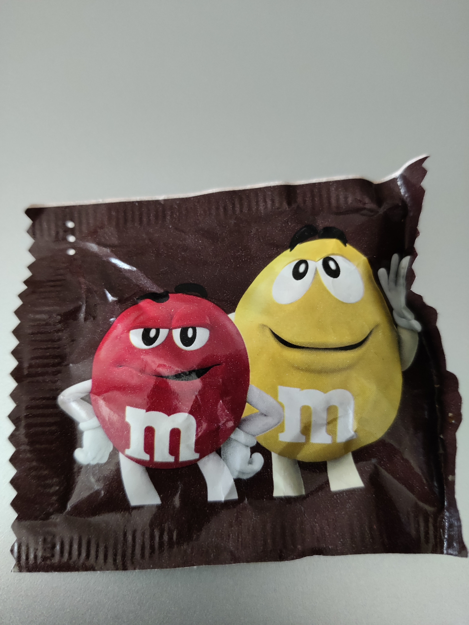 The image shows a close-up of a dark brown M&M's candy wrapper against a light grey background. The wrapper features two M&M character illustrations: a red M&M on the left and a yellow M&M on the right. Both characters have white arms and the letter "m" printed on their bodies in white. The red M&M has a smug expression with its eyes narrowed and a slight smile, while the yellow M&M has wide eyes and a cheerful smile, with its right hand raised as if waving. The wrapper is slightly wrinkled and torn at the top right corner. The scene does not provide enough information to determine its location within Lower Paxton Township, United States of America.