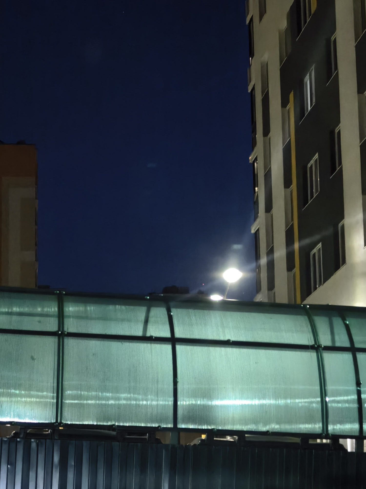 An exterior, ground-level shot of a modern building at night. The building is tall and appears to be under construction or recently built, with a mix of dark gray and cream-colored panels and numerous windows. A bright, artificial light illuminates the scene from the left, casting a glow on the building and a translucent, green-tinted canopy in the foreground. The sky above is a deep, dark blue. In the distance, a faint light is visible in the sky, possibly a star or a distant airplane. A dark, ribbed metal fence is at the bottom of the frame, partially obscuring the view of the ground.