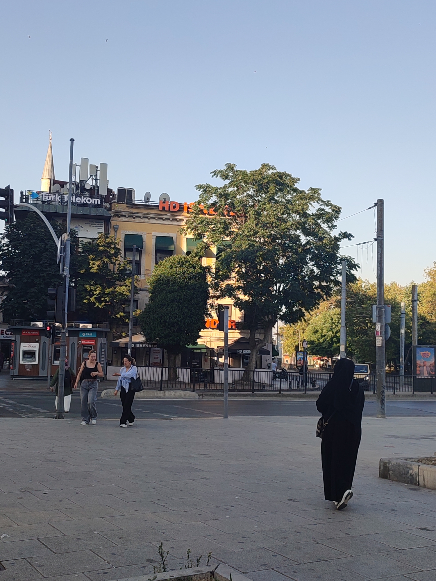 In Kadıköy, Turkey, the image captures a street scene with a building bearing a "Turk Telekom" sign and an orange "HD Iskender" sign. Several individuals are present: a person in a black abaya walks away from the camera towards the right; two women, one in a black tank top and grey pants, and the other in a light blue shirt and dark pants, walk towards the left on a sidewalk; and another person in a green headscarf and white garment walks from right to left, partially obscured. An ATM is visible on the left side of the frame. The environment includes trees, streetlights, power lines, and a clear sky. The ground is paved with grey tiles.