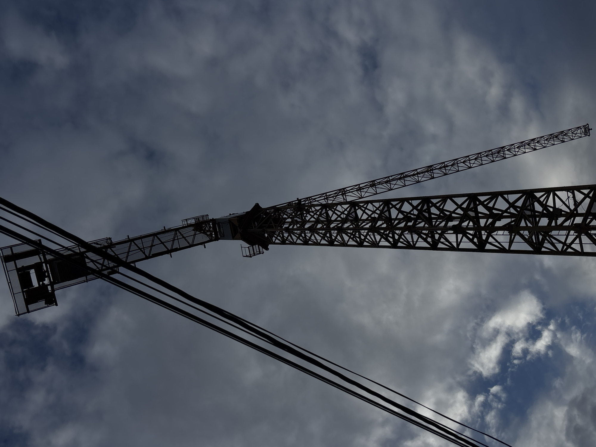 A low-angle shot captures a construction crane silhouetted against a cloudy sky in Belgrade, Serbia. The crane's lattice boom stretches diagonally across the frame, with a counterweight structure on the left. Three electrical power lines intersect the image in the lower-left corner. The sky features a mix of dark gray and patches of blue, suggesting overcast conditions.
