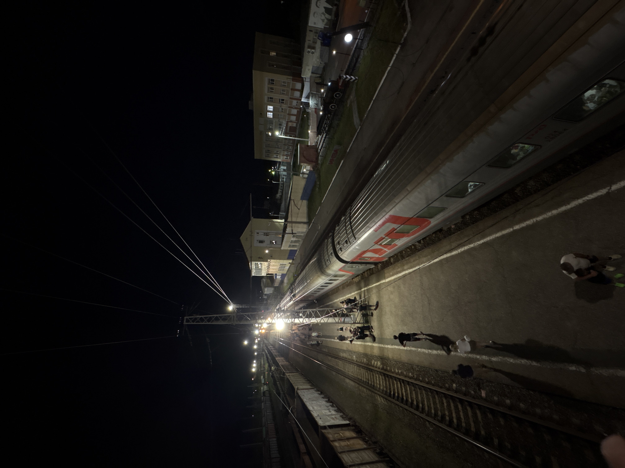 This is an aerial, nighttime, long-shot photograph taken from above a train station platform. The scene depicts a train stopped at the platform, with several people milling about.
In the foreground, the train car is visible, with its windows reflecting the ambient lights. The train appears to be red and grey. A few people are standing on the platform near the train, some walking and others gathered in small groups. One person in the lower right corner is wearing a white t-shirt and green sneakers.
The platform itself is paved and appears to be illuminated by overhead lights and lights from the train. Adjacent to the platform, train tracks run parallel, with several freight cars visible on the left side of the image. Further in the background, buildings line the side of the tracks, appearing to be station buildings or other industrial structures. Overhead power lines are strung across the upper portion of the image, supported by utility poles. The sky is dark, indicating nighttime.
The location is the Inza train station in Inza, Russia.