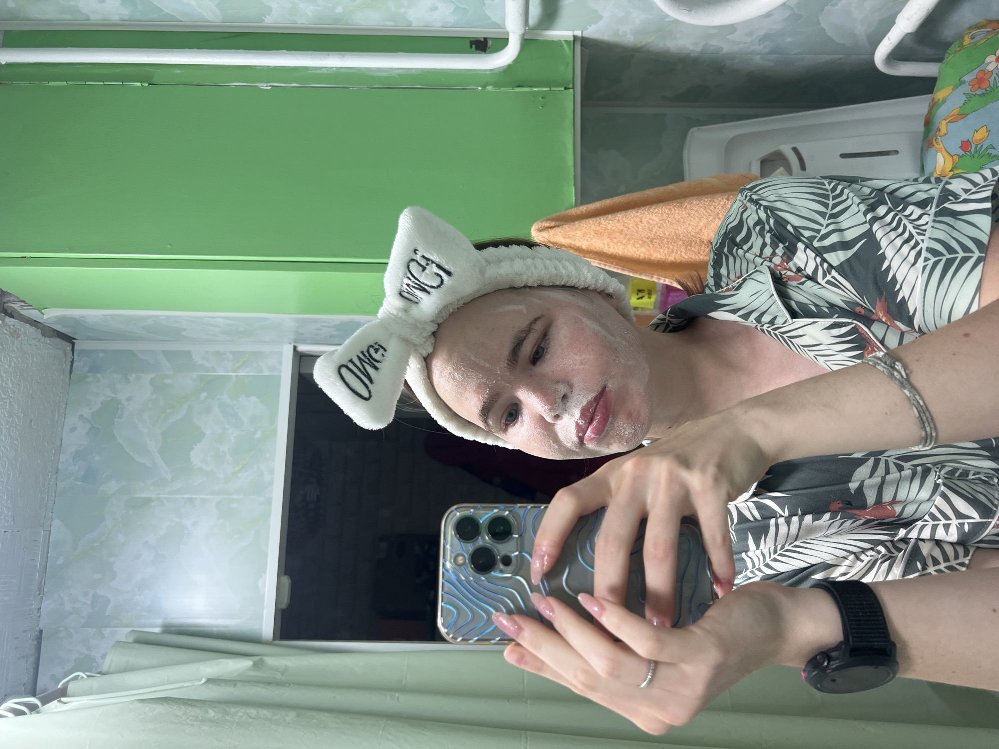 The image shows a young woman with a white fluffy headband with the word "OHMY" embroidered on it in black letters, and a facial mask on her face. She is holding a smartphone in her left hand and taking a selfie in a bathroom. The woman is wearing a pajama top with a tropical leaf print and a pink flamingo design. She has long, manicured nails with a light pink polish. A silver ring is visible on her left ring finger, and a silver bracelet is on her right wrist. A black smartwatch is on her left wrist. The bathroom has light green tiled walls and a green cabinet. An orange towel is hanging on the right side of the cabinet. The environment appears to be a residential bathroom. This image was taken in Chaikovsky, Russia.