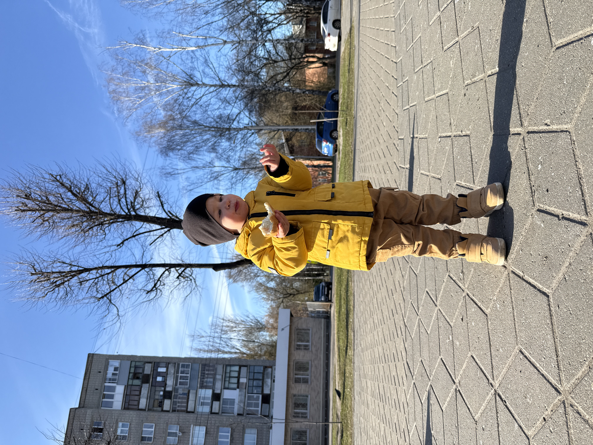 A young child stands on a paved walkway. The child is wearing a yellow jacket, brown pants, and a dark beanie. The child is holding something in their left hand. The walkway is made of gray pavement stones with a diamond pattern. The sky is clear and blue with bare trees visible in the background. Behind the trees are buildings, including a multi-story apartment building with mismatched windows. Parked vehicles are visible along the street. The location appears to be Rusanovo, Russia.