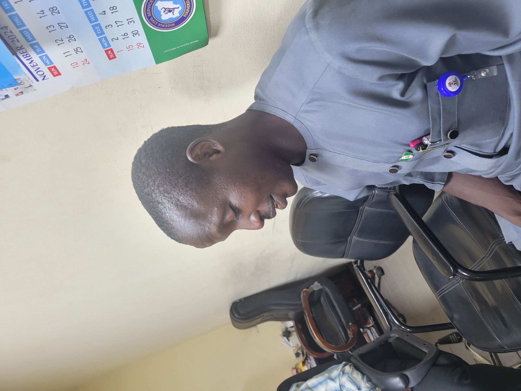 The image shows a man in a gray traditional outfit, possibly a kaftan, seated in an office environment. He is looking downwards and to his right. On his chest, he has a pocket with a few items clipped to it, including pens and a lanyard with a blue retractable reel attached. Behind him, there are several black leather office chairs, some appearing to be unoccupied. To his right, a wall with a calendar is visible, indicating "NOVEMBER 2024" and showing a grid of dates and days of the week. A circular logo with text that appears to say "NIGERIA CIVIL SERVICE" is also visible on the calendar. The lighting suggests an indoor setting, likely an office. The presence of the "Nigeria Civil Service" logo strongly suggests the location is within an office related to this organization in Abuja, Nigeria.