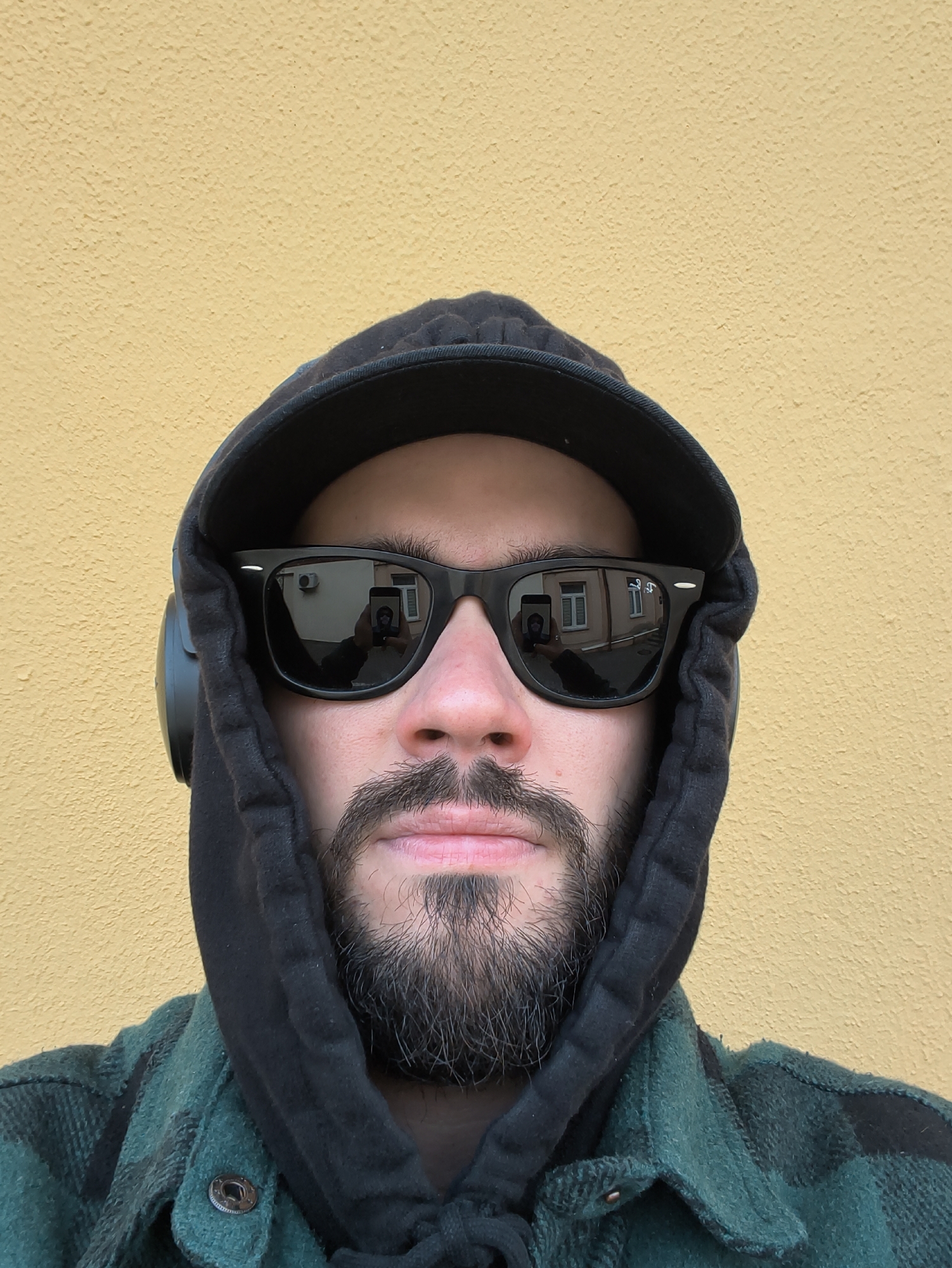 A male individual is depicted in a close-up, head-and-shoulders portrait against a textured, plain light-yellow wall. He wears black Wayfarer-style sunglasses, a black hooded sweatshirt with the hood drawn up, and a black baseball cap underneath the hood. A green and black plaid flannel shirt is visible below the sweatshirt. Black over-ear headphones are worn on his left ear. The subject has a dark beard and mustache. The reflections in the sunglasses indicate the scene is an urban street within Hrodna, Belarus. The left lens reflects a pale yellow/cream-colored building with a white-framed window and a rectangular air conditioning unit mounted above it. The right lens reflects a light-colored (beige/light brown) multi-story building featuring prominent white-framed, multi-pane windows and a paved street area. Both reflections also show the subject's arm holding a dark smartphone, confirming the image is a selfie.
