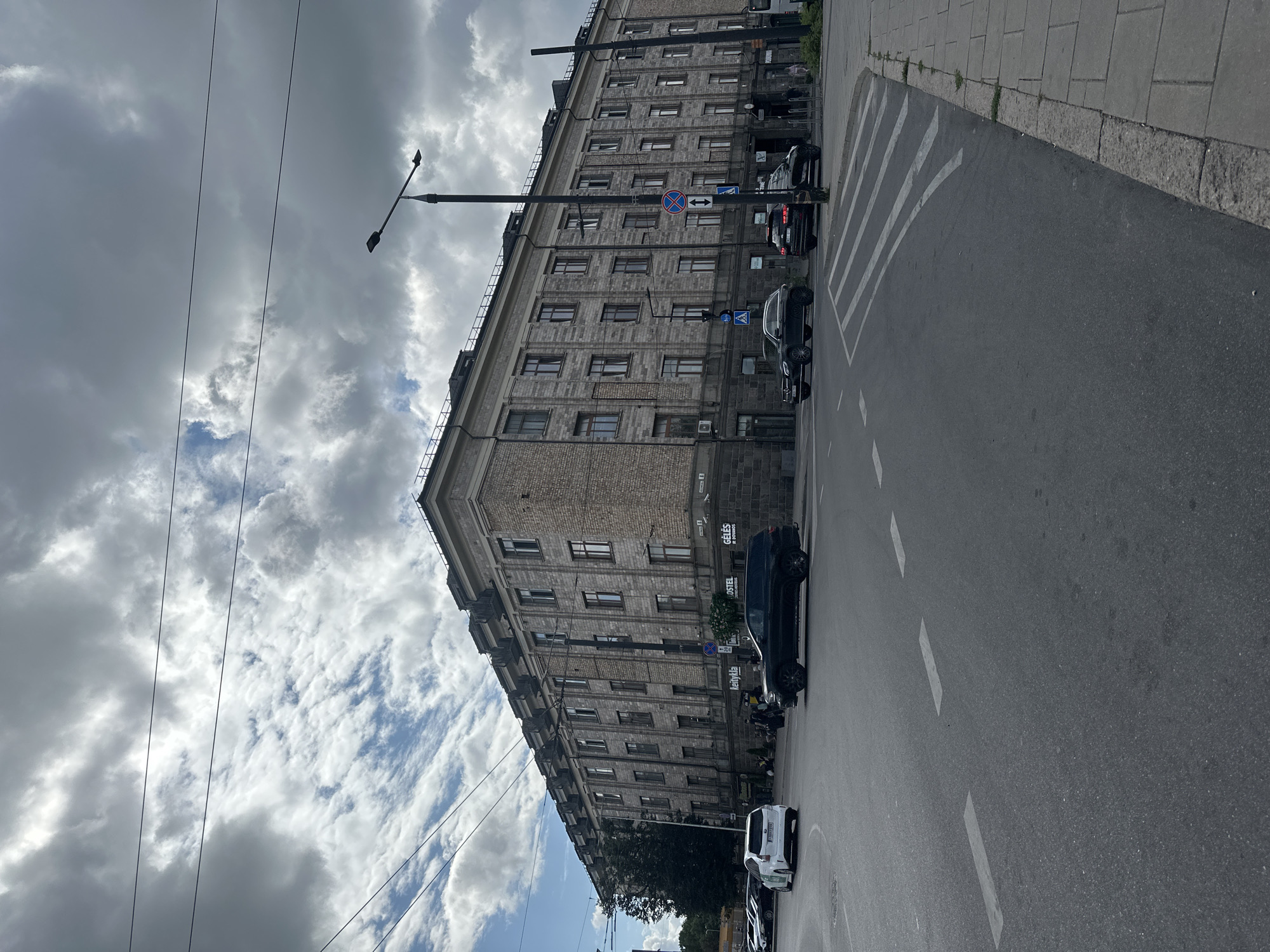 An eye-level, medium shot captures a grand, multi-story building in Vilnius, Lithuania, under a cloudy sky. The building, constructed with grey stone and brick, features numerous windows and decorative elements along its facade. In the foreground, a paved intersection with visible white lane markings and a pedestrian crossing is present. Several cars are on the road, including a white hybrid sedan, a black SUV, and other dark-colored vehicles. A tall street lamp stands near the intersection. Pedestrians are visible on the sidewalk in front of the building. The scene appears to be a typical urban streetscape with a mix of architectural styles and contemporary traffic.