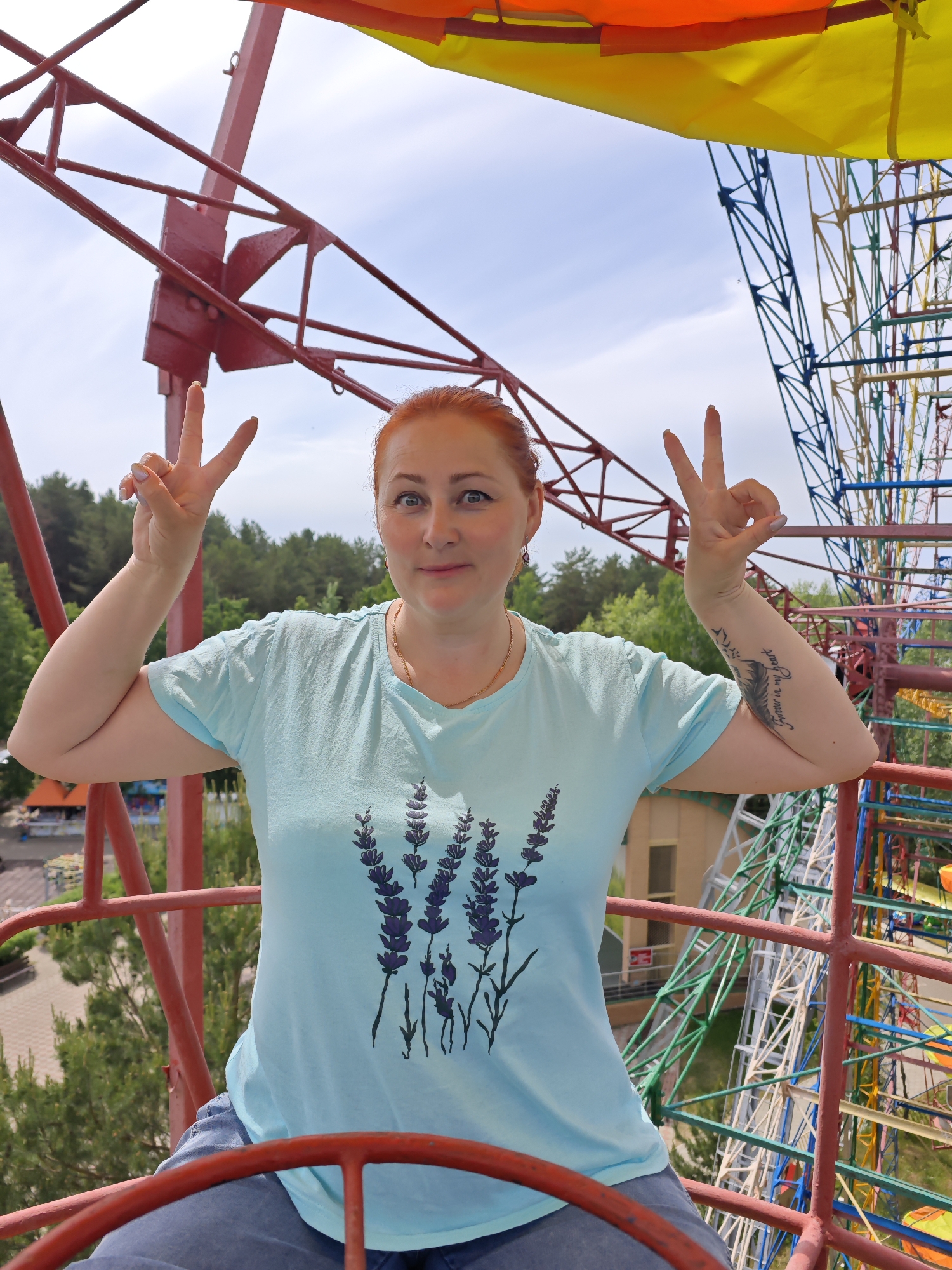 A woman with red hair is making the peace sign with both hands while sitting on a red, iron structure, likely the steering wheel of a Ferris wheel car, located in Chaikovsky, Russia. The woman wears a light blue shirt with a floral lavender print and has a tattoo on her left arm. A colorful Ferris wheel and green trees are visible in the background, along with a building. The sky is partially cloudy.