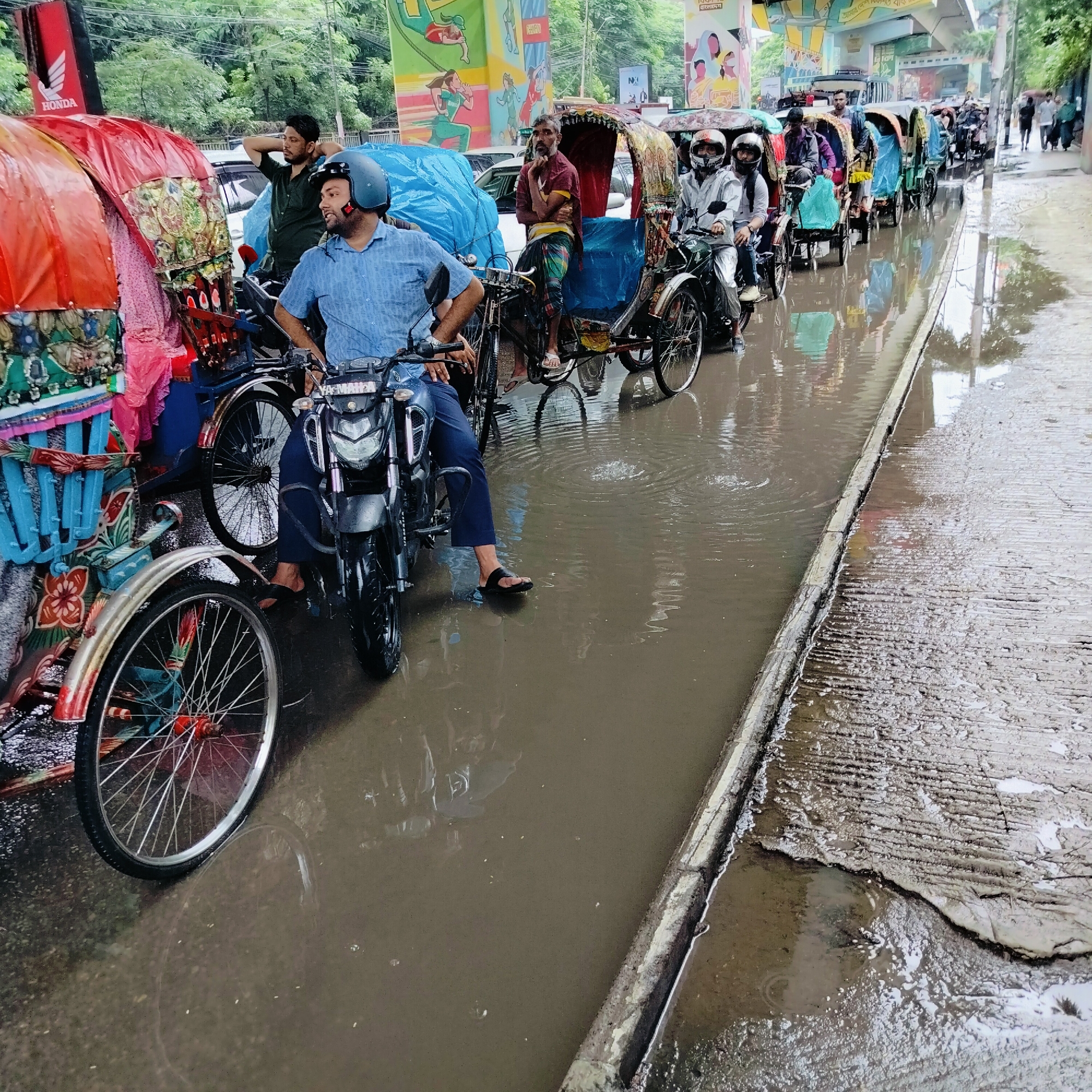 The image captures a scene on a flooded street in Dhaka, Bangladesh, likely during or after rainfall, as evidenced by the standing water and reflections on the road. In the foreground, a man wearing a blue shirt and a helmet is seated on a motorcycle. Next to him, a colorful cycle rickshaw, a common mode of transport in Dhaka, is parked. The rickshaw is adorned with intricate designs and brightly colored fabrics. Further down the street, a line of other motorcycles and cycle rickshaws are visible, some carrying passengers. People are seen in various positions, some riding, others waiting. The background reveals a busy urban environment with trees, buildings, and billboards, one of which features a "Honda" logo. The overall atmosphere is one of urban life continuing amidst challenging weather conditions.