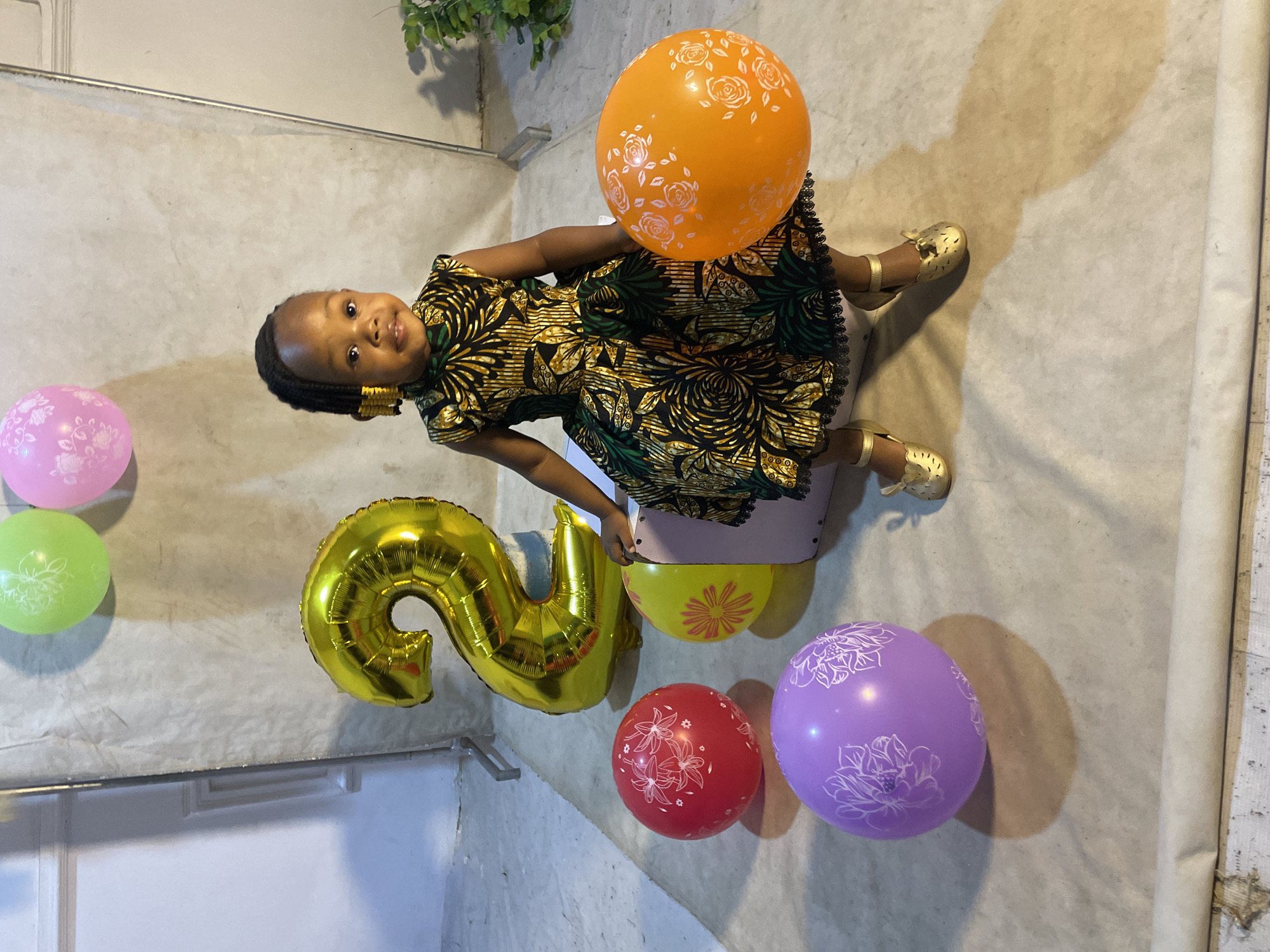 A young girl with braided hair and gold accents on her hair, wearing a colorful dress and gold shoes, sits on the floor surrounded by balloons. She holds an orange balloon with white floral patterns in her right hand, while a large gold balloon shaped like the number "2" is behind her. Several other balloons, including red, purple, and yellow, are scattered around her. The background consists of a textured backdrop and a hint of a potted plant. The lighting suggests an indoor setting, possibly a home studio, given the seamless backdrop and controlled lighting. The scene is indicative of a birthday celebration, likely for the girl's second birthday. The specific location within Jos, Nigeria, cannot be determined from the visual information provided in the image.