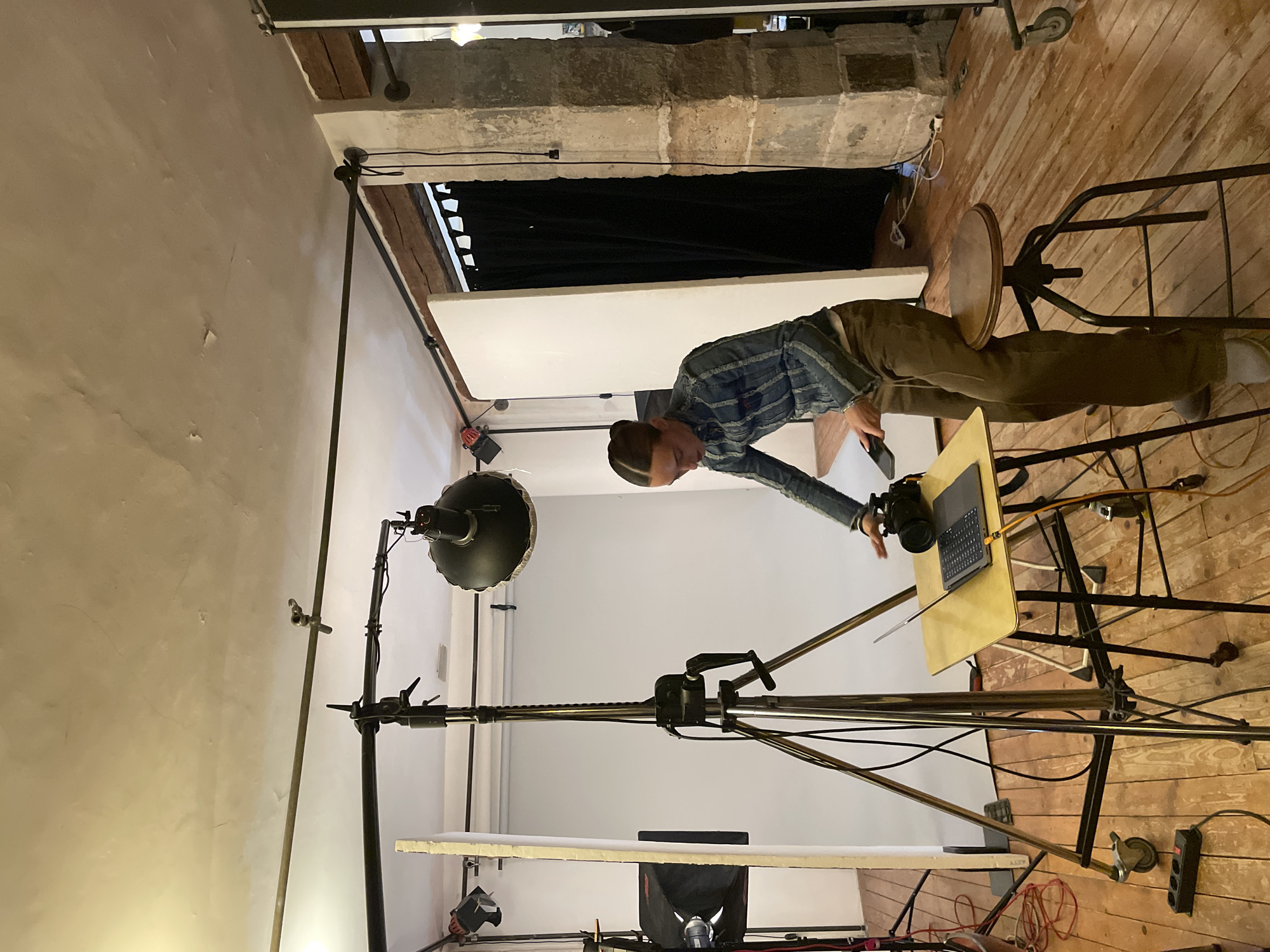 The image depicts an indoor photographic studio setting located within an old building in Paris, France. A young adult individual, with hair pulled back, is observed leaning over a small, wheeled table. They are dressed in a blue striped jacket, a light undershirt, and brown trousers. The individual's right hand is positioned on a black camera tethered via a yellow cable to an open silver laptop also on the table. A black smartphone is held in their left hand.
The environment features an unpainted, textured ceiling showing cracks, and distressed wooden floorboards. An exposed stone pillar and a rough wooden beam are visible on the right side of the frame, indicating the age of the structure. White photography backdrops or reflector panels are set up against walls, and black curtains are visible in a doorway opening on the right.
Photography equipment is prominent, including a large, chrome-plated light stand with a crank mechanism in the center, and two studio lights—one with a softbox and another with a parabolic reflector—mounted on additional stands. Overhead, metal pipes are rigged across the ceiling, likely part of an equipment grid. Various black and orange cables are scattered across the floor, connecting to a black power strip. A dark wooden stool with metal legs is situated near the individual. The scene suggests an active photo or video production setup.