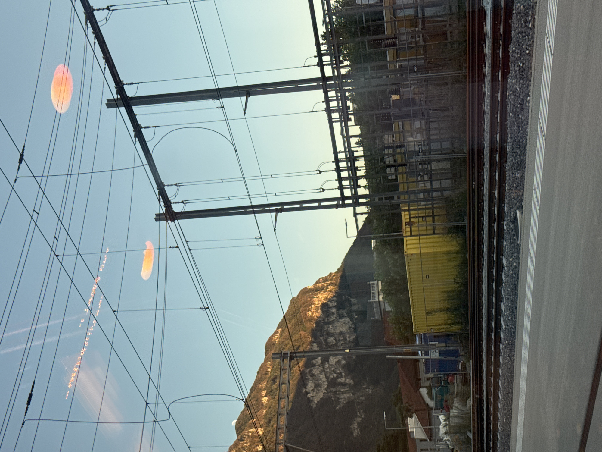 The image shows a view of the sky above a train track and a mountain range. The sky is clear with a few wispy clouds and two lens flares, one large and orange, the other smaller and oval shaped and orange. There are numerous overhead power lines and poles. The mountain range is steep and rocky, with some trees on its lower slopes. The sunlight illuminates the side of the mountain, casting shadows. Below the mountain are some buildings and a yellow shipping container. In the foreground are train tracks and a paved area. The scene appears to be in the city of Val Mara, Switzerland, possibly near a train station.