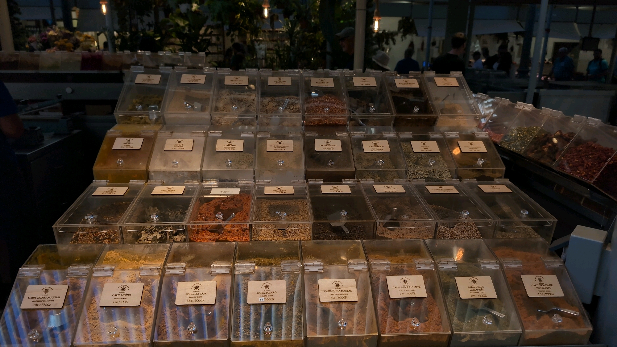 The image depicts a marketplace stall in Porto, Portugal, featuring a variety of dried spices displayed in clear, hinged plastic bins. The bins are arranged in tiered rows, with labels indicating different types of curry and spices, such as "CURRY INDIA ORIGINAL," "CURRY MOLIDO," "CURRY LONDON," "CURRY BOLHÃO," "CURRY INDIA MADRAS," "CURRY INDIA PICANTE," "CURRY VERDE TAILANDES," and "CURRY VERMELHO." Some bins contain loose spices, while others hold whole or ground ingredients. Small scoops or spatulas are visible in some of the containers.
In the background, blurred figures of people are present, suggesting a bustling market environment. The lighting appears to be artificial, with spotlights illuminating the products. The overall scene conveys a commercial setting focused on the sale of spices. The stall appears to be part of a larger market or shop, with additional shelving and products visible in the obscured background. The wooden labels on the bins are consistent with a European style of product labeling.