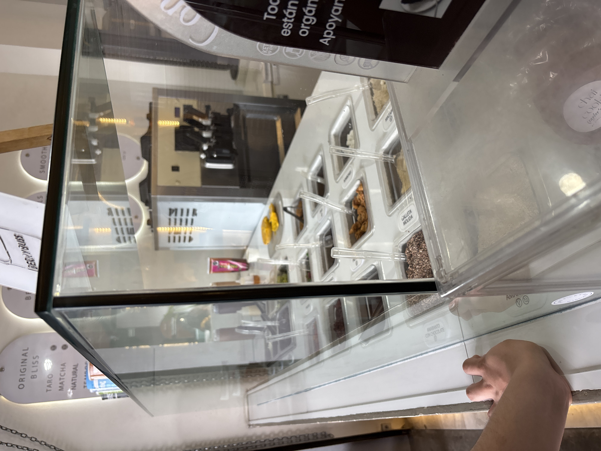 The image shows the inside of a store or café in San Bernardino Tlaxcalancingo, Mexico, likely a yogurt or smoothie shop. A person's hand is visible in the foreground, near a clear plastic container labeled "AMARANTO CON CHOCOLATE" and another container with a "chai cook" label. The containers appear to hold toppings or ingredients for the yogurt/smoothies. Behind the containers are a variety of other toppings. In the background is a display of menu options like "ORIGINAL BLISS," "TARO," "MATCHA," "NATURAL" and "BERRY BLISS." Also there is an ice cream machine and other kitchen equipment.