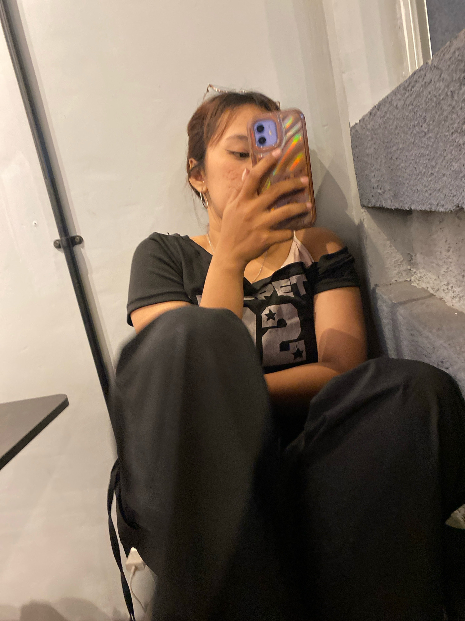 A young woman with brown hair and freckles is sitting on a concrete staircase, holding up a phone to take a selfie. She is wearing a black off-the-shoulder shirt with a graphic design and black pants. The background is a plain gray wall with a black pole running vertically along it. The scene appears to be outdoors, possibly in a public space within Klurak, Indonesia.