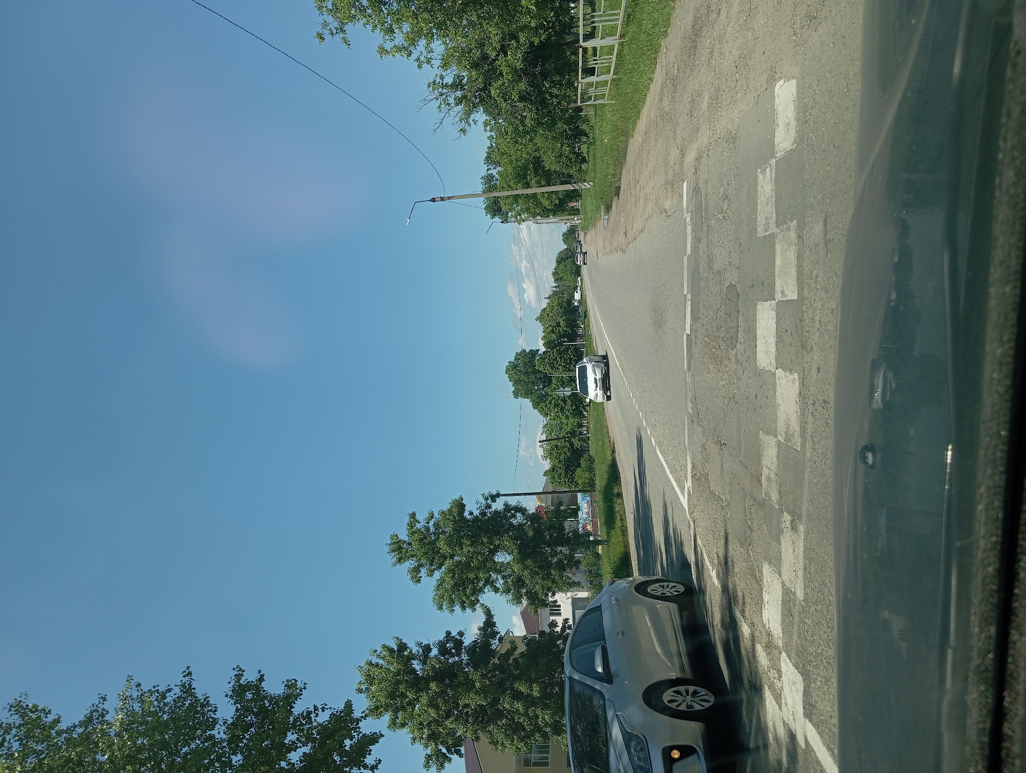 A street scene in Otradnaya, Russia, under a clear blue sky. The asphalt road features a pedestrian crossing marked with white squares. On the left side of the road, a silver sedan is parked, its front wheel near the crossing. Further down the road, other vehicles are visible in the distance. Trees line both sides of the road, and buildings can be seen behind the trees on the left. A metal fence runs along the right side of the road. The overall scene depicts a typical day in a Russian town.